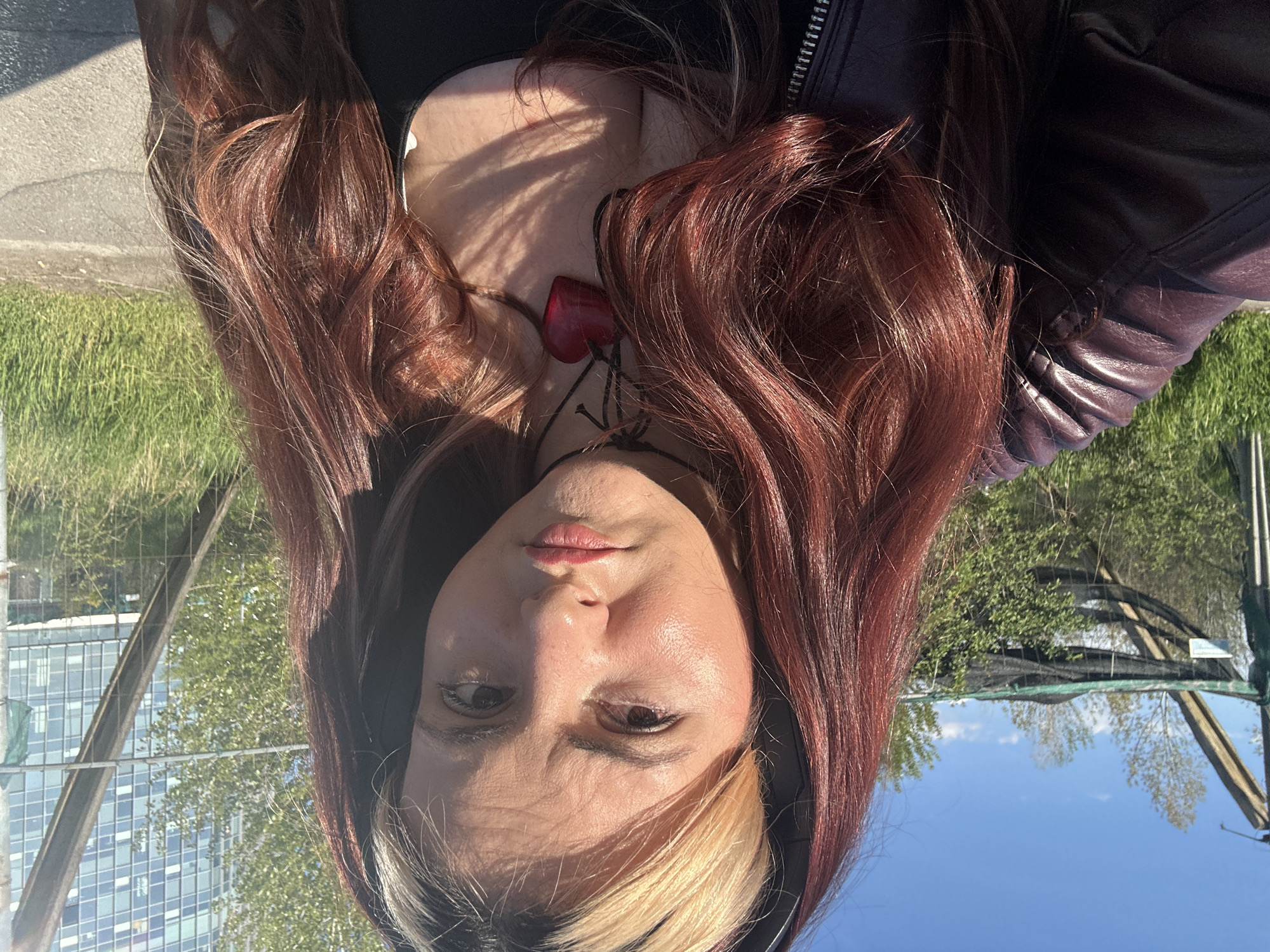 The media file features a close-up portrait of a person, captured outdoors during the daytime. The subject has long, wavy hair, predominantly reddish-brown, with a distinct blonde and white streak on the left side of her face. She has a fair complexion and a neutral expression, looking directly into the camera. She is wearing a dark top, possibly a tank top, beneath a dark, zippered jacket, likely made of leather. A prominent red heart-shaped pendant on a dark cord necklace rests on her chest.
The bright sunlight casts noticeable shadows, particularly from her hair onto her chest and face, indicating a sunny day. The background features a mix of urban and natural elements. To the left, a paved surface runs alongside green grass. To the right, a clear blue sky with sparse white clouds is visible. In the upper background, there are green trees, modern buildings with numerous windows, and metallic structures, possibly scaffolding or part of a fence. The overall setting suggests an outdoor urban or park environment, within Sofia, Bulgaria. No text is visible in the image.