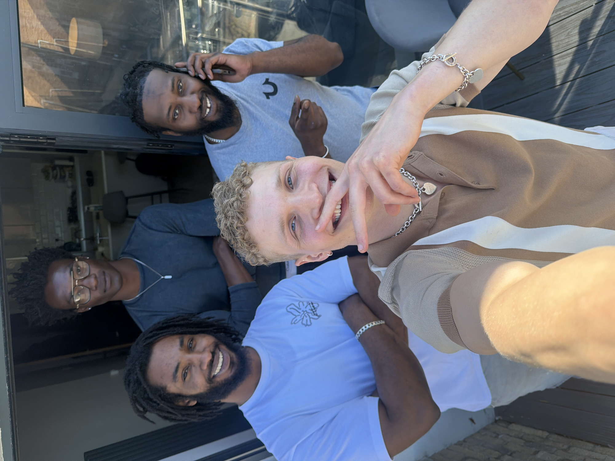 The image captures a vibrant group selfie of four men outdoors on a sunny day, likely on a patio or deck area with visible wooden flooring. In the foreground, on the right, a man with light skin and curly blonde hair is taking the photo, smiling broadly. He wears a striped brown and white polo shirt, a silver chain necklace with a "Tiffany & Co." pendant, and a matching silver bracelet with heart-shaped charms.
Behind him, to the left, another man with dark skin and dreadlocks is smiling while holding a phone to his ear. He is wearing a light blue t-shirt with the distinctive "True Religion" horseshoe logo. Further back, on the far left, a third man with dark curly hair and glasses looks towards the camera, dressed in a dark grey t-shirt. In the lower left section of the frame, a fourth man with dreadlocks and a beard is also smiling widely, wearing a white t-shirt with a grey abstract or floral graphic design and a silver bracelet.
The bright daylight casts distinct shadows, suggesting it is midday or early afternoon. A modern glass door or window behind the men reflects the surroundings and reveals a blurry interior space. All the individuals are smiling, conveying a mood of happiness and camaraderie in this outdoor setting, consistent with a casual gathering in London, United Kingdom.