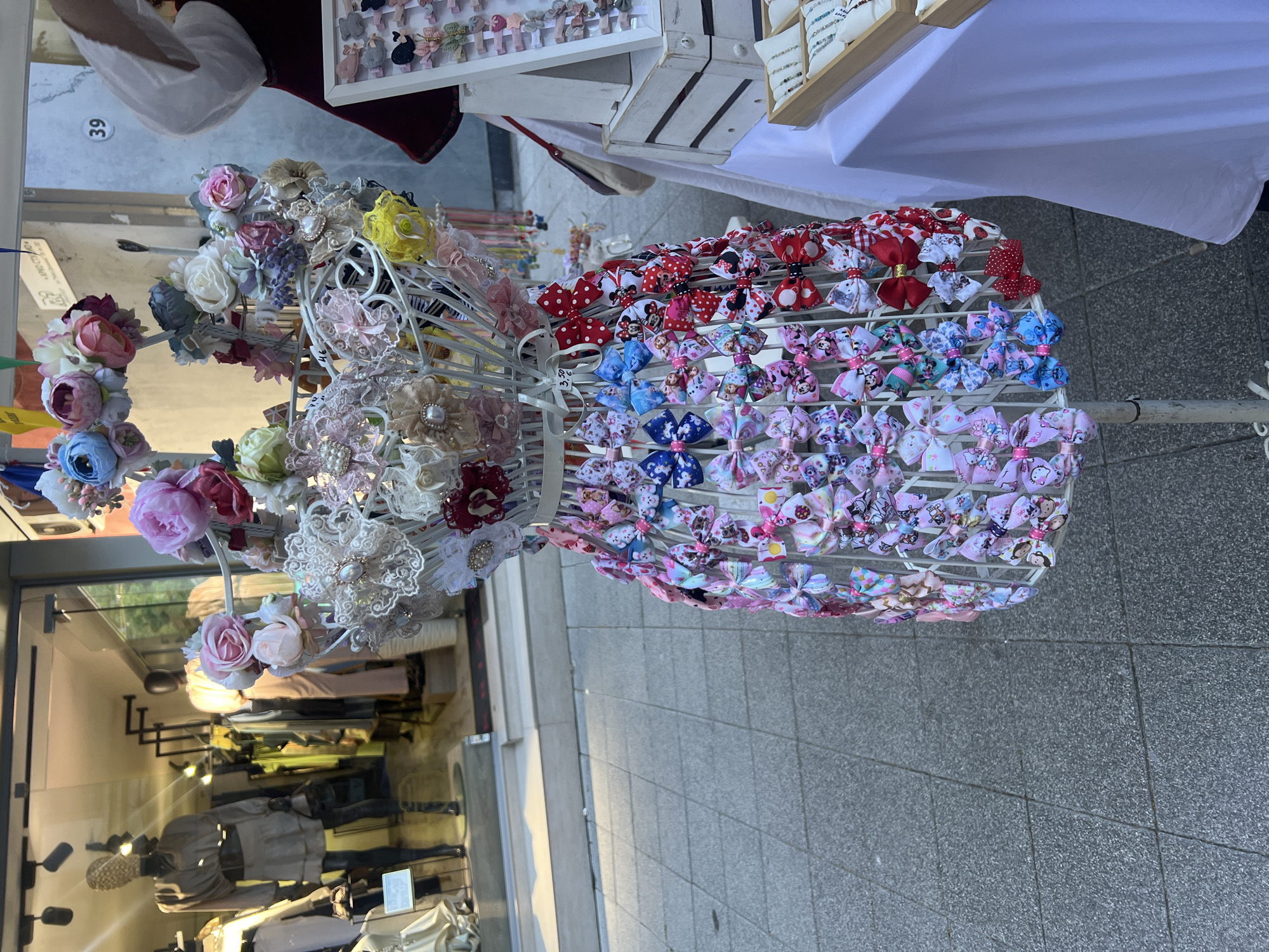 The image shows a display stand of hair accessories, likely outdoors in a commercial area. The stand is metal and painted white. It holds various hairbands and hair bows. The hairbands are decorated with fabric flowers and lace. The hair bows are patterned with characters such as Minnie Mouse, Frozen, and Hello Kitty. A shop window is visible in the background, reflecting the items on the display stand and the surrounding area. Mannequins are visible in the shop. The ground is paved. Based on the architectural style and general appearance, the location is possibly in Plovdiv, Bulgaria.