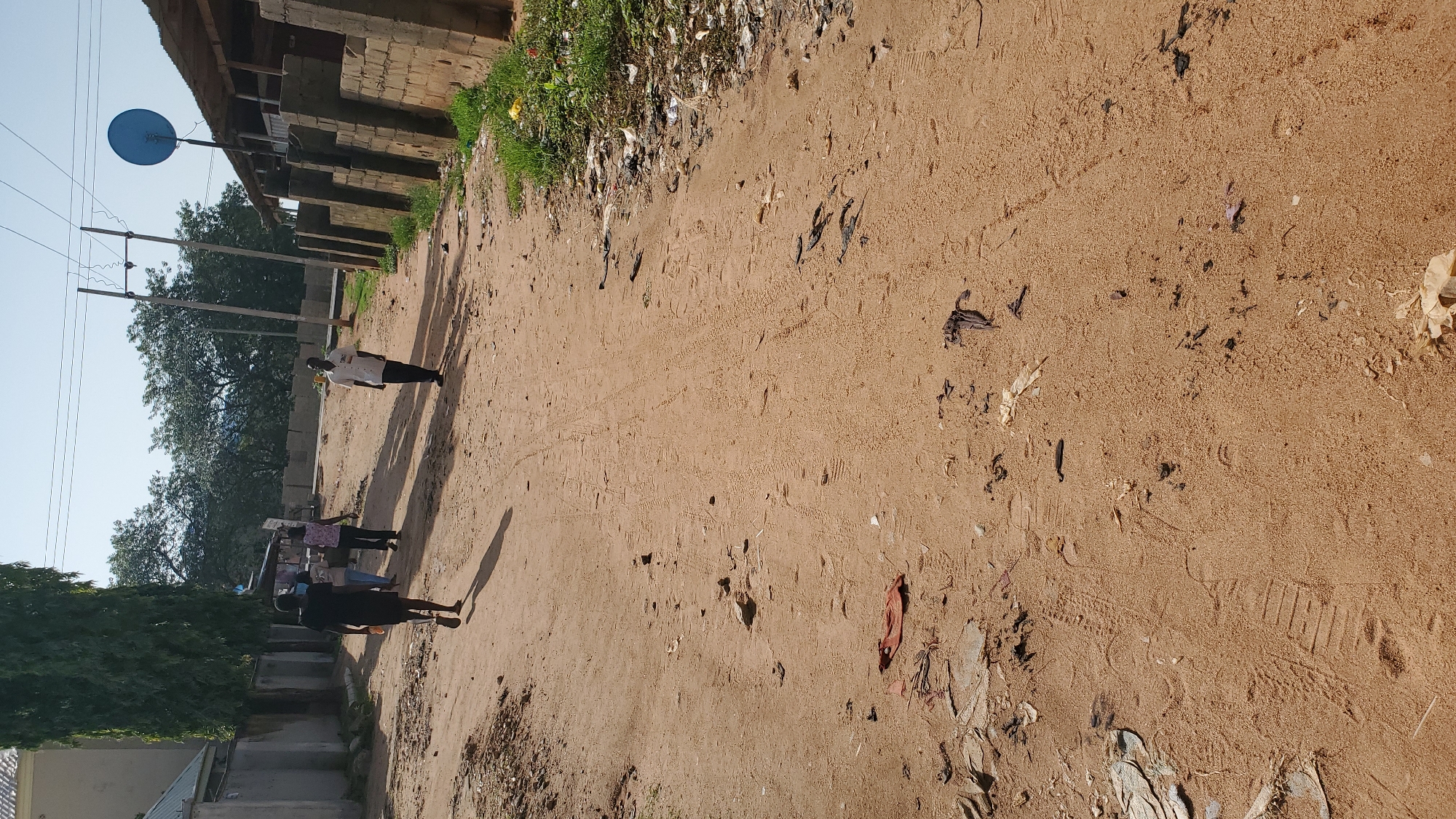 In Jalingo, Nigeria, a dirt road traverses a neighborhood, with a cluster of buildings on either side. The road is unpaved, marked by tire tracks and scattered debris. A few individuals are walking on the road, some heading towards the foreground and others in the distance. On the right side of the road, a structure with a corrugated metal roof stands, built with brick walls. A blue satellite dish is mounted on a pole near this structure. The area appears to be in a state of disrepair, with visible litter along the roadside and on the ground. The sky is clear and bright, indicating daylight.
