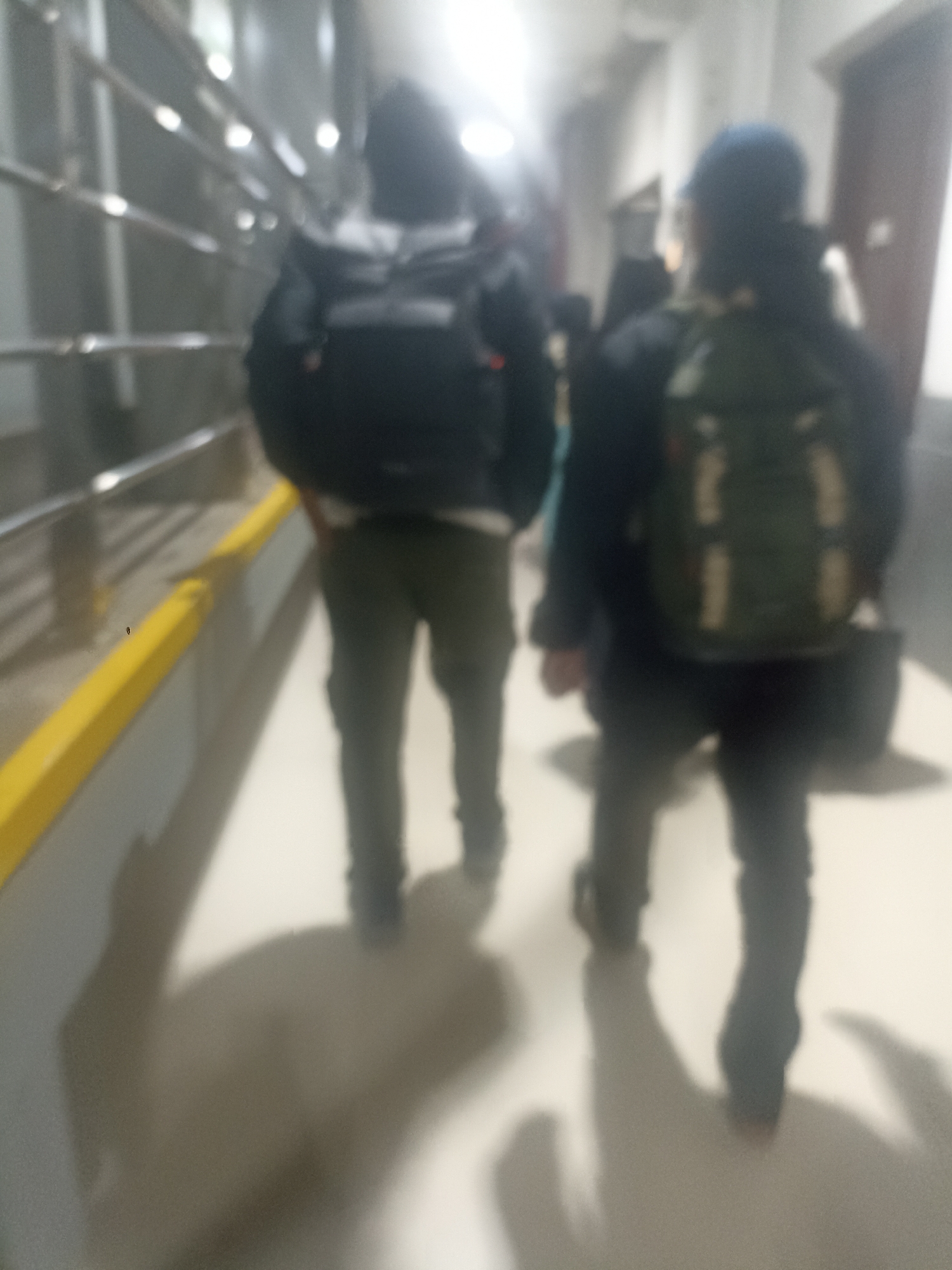 The image shows two individuals walking down a corridor, possibly a train station or an airport, with backpacks on. The environment is dimly lit, with fluorescent lights casting reflections on the metallic handrail and the floor. The person on the left is wearing a dark jacket and olive-colored pants, carrying a black backpack. The person on the right is wearing a dark jacket and dark pants, with a green backpack and a blue cap. Their faces are not clearly visible due to the blurriness of the image. The corridor appears to be clean and well-maintained. The scene is devoid of any specific identifiable landmarks that would place it in Bandung City, Indonesia.
