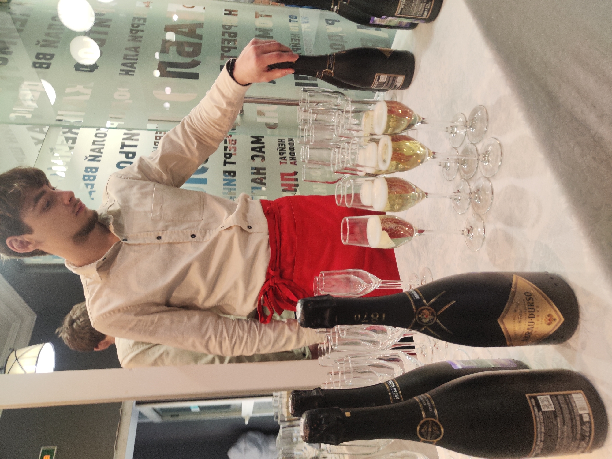 The image depicts a vertical shot of a celebratory scene, likely a party or event, with a focus on bottles of sparkling wine and glasses. A young man, wearing a light-colored button-down shirt and a red apron tied at the waist, is in the process of pouring wine from a dark bottle into a row of waiting champagne flutes. His arm and hand are prominent as he performs the pouring action.
In the background, there is a glass wall or partition with reflective qualities, showing blurred text that appears to be in Russian, indicating the location is Saint Petersburg, Russia. The text "ABRAU-DURSO", "1870", and "SPARKLING WINE" are visible on the bottles, identifying the brand of the beverage.
The setting appears to be indoors, possibly a restaurant, bar, or event hall, with a white tablecloth laid out on a surface where the bottles and glasses are arranged. The lighting suggests it could be evening or a dimly lit interior space. The overall mood conveyed is one of anticipation and festivity, with the poured wine creating a sense of effervescence and celebration. Several bottles of the sparkling wine are lined up, ready to be served, and multiple glasses are already filled with the golden liquid.