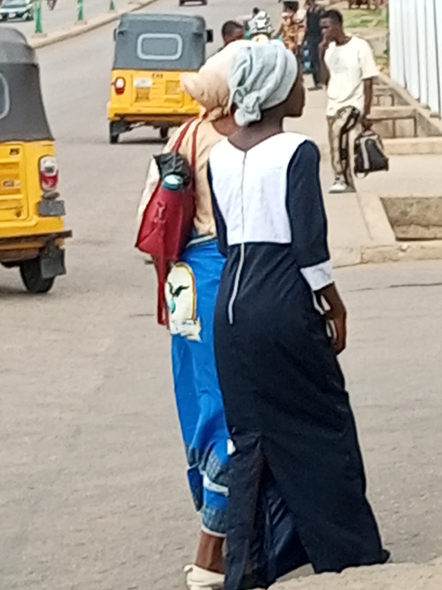 In Gombe, Nigeria, two individuals are walking on a paved road next to a sidewalk. The person in the foreground is wearing a navy blue, full-length dress with white trim on the sleeves and a white panel on the chest. They have a headscarf on. Behind them, another person is wearing a blue, full-length dress with white trim and a headscarf. They are carrying a red bag with a white design on it. In the background, a yellow motorized tricycle is parked on the side of the road. Further in the background, there are more people, some walking and some standing, and another yellow motorized tricycle. The environment appears to be a typical urban street scene in Nigeria.
