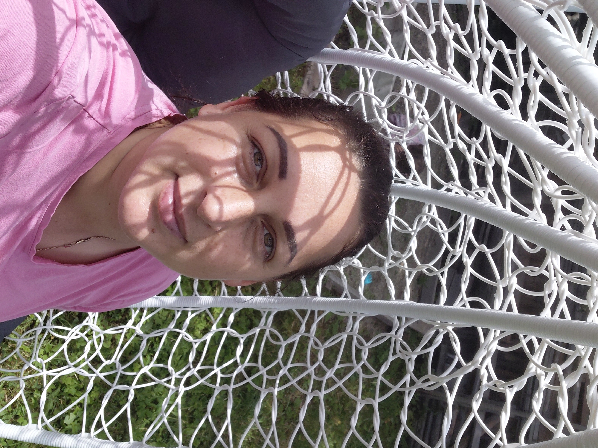 In Chernihiv, Ukraine, a woman with dark hair and light green eyes is captured in a selfie from a low angle. She is wearing a pink t-shirt and is seated inside a white wicker chair with a net-like pattern. The chair is outdoors, as evidenced by the grassy ground visible through the netting. Sunlight casts dappled shadows across her face, highlighting her features. To her left, a dark gray sleeve is partially visible, suggesting she is not alone, or at least is wearing multiple layers. The background shows a wooden fence and more greenery. The overall scene suggests a casual, outdoor setting.