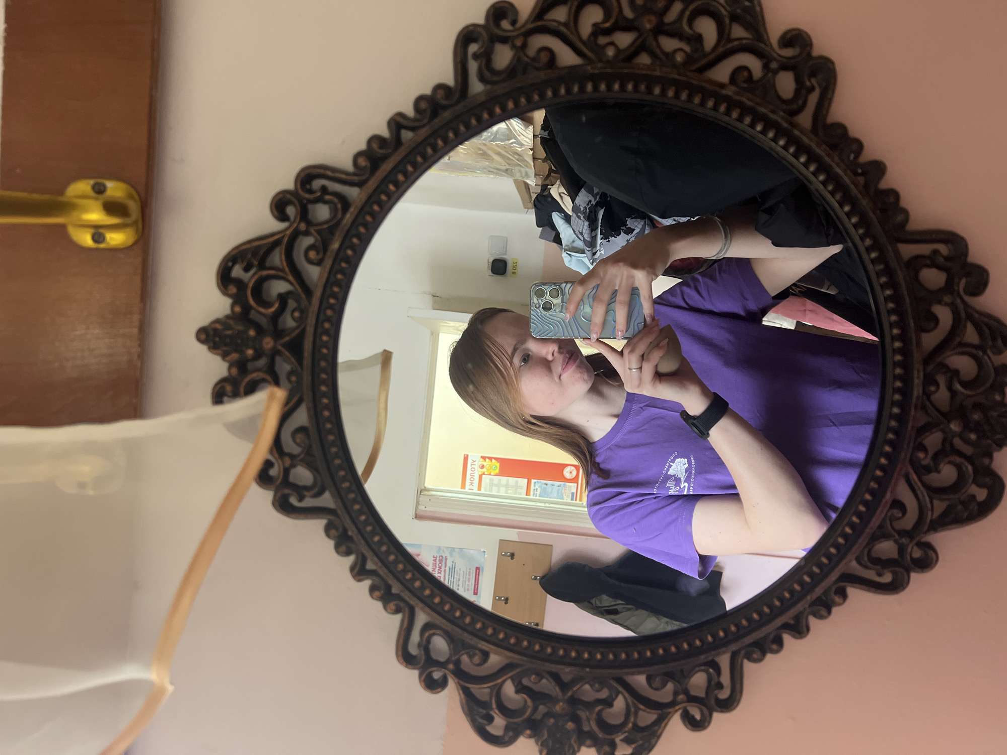 A young woman is taking a selfie in a decorative, round, dark-toned mirror. She is wearing a purple t-shirt and a dark watch on her left wrist. She is holding a light-colored cell phone with her right hand, presumably to take the photo. The reflection in the mirror shows a room with a window, posters on the wall (with text in a non-English script), and various items of clothing scattered around. A golden door handle is visible on a wooden door to the left of the mirror. The environment appears to be a room with pinkish walls. Based on the text on the posters, the location is Berezniki, Russia.
