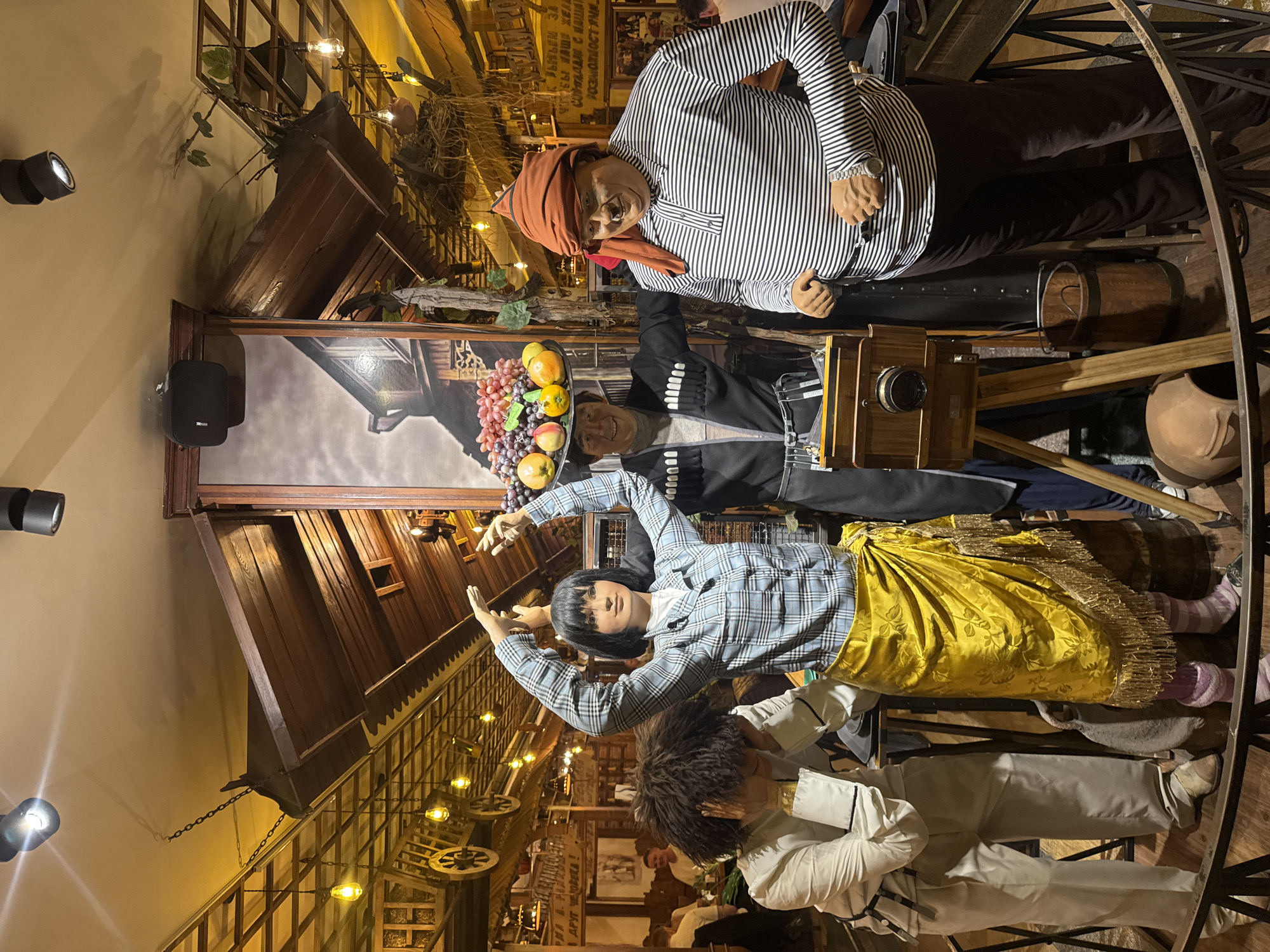 The image shows a staged scene inside a restaurant in Yerevan, Armenia, featuring a historical depiction of Armenian life with mannequins dressed in traditional clothing. A man in a striped shirt and a red turban stands near a wooden camera. A man wears a chokha with a tray of fruit on his head. A woman with black hair wears a patterned shirt and a yellow skirt and has her arms raised. A man wearing a white shirt and pants stands in front of her. The restaurant has wooden beams and ornate lighting.