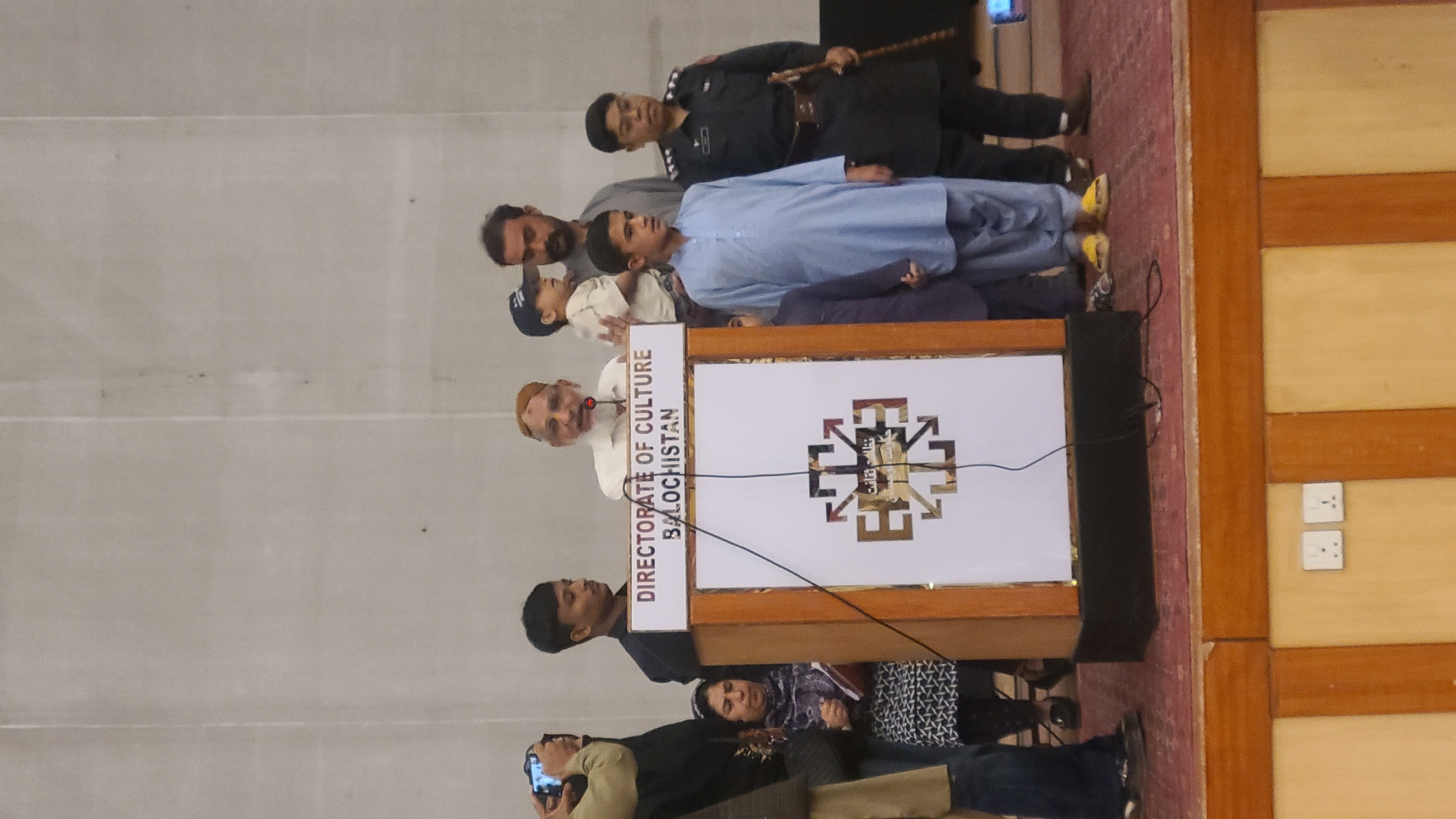 A medium indoor shot captures a man speaking at a podium in Quetta City Tehsil, Pakistan. The podium reads "Directorate of Culture Balochistan." A logo featuring a geometric design is on the lower half of the podium. Several people stand behind the speaker, including a boy in a light blue shirt and a man in a dark uniform. Some audience members sit in the foreground. The room has a wall with electrical outlets.