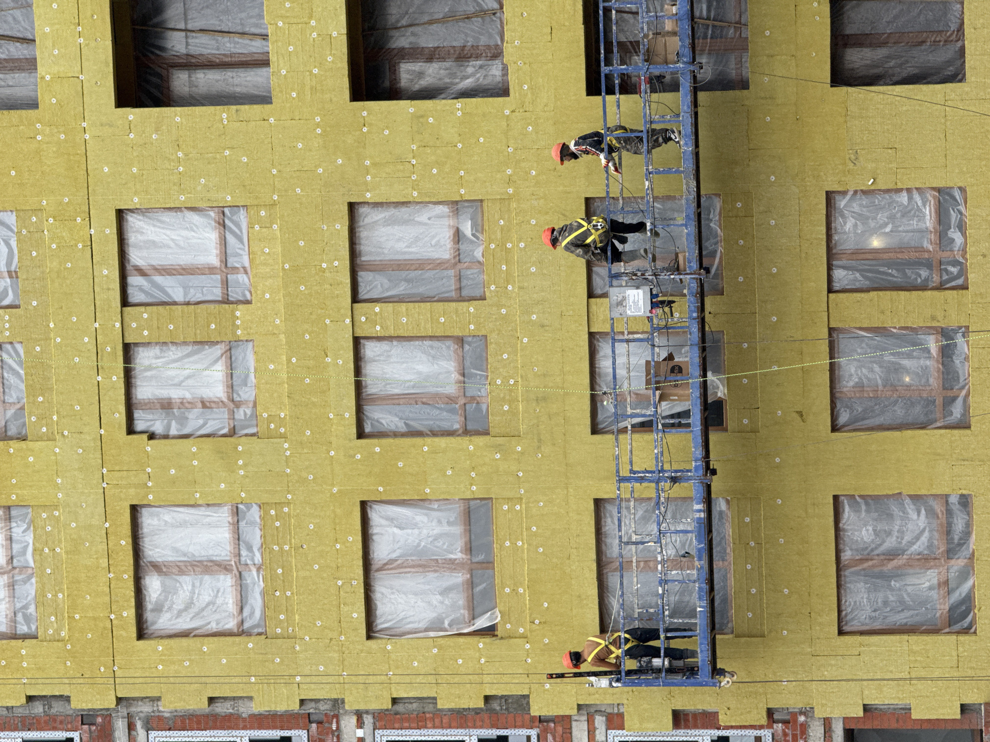 The facade of a multi-story building undergoing construction in Perm, Russia, is depicted. The dominant surface consists of yellow-green mineral wool insulation panels, uniformly secured by numerous white disc fasteners. Multiple rectangular window openings reveal installed wooden frames, each covered with translucent plastic sheeting. A blue suspended work platform, featuring a metal railing, is positioned mid-frame, approximately at the building's sixth-floor level. Three individuals, identified as construction workers, are present on the platform. All three wear red hard hats and yellow safety harnesses. The leftmost worker stands, holding a yellow spirit level against the insulated facade. The central worker is seated on the platform's edge. The rightmost worker stands, holding a tool or piece of equipment. Exposed red brickwork and a white-framed window are visible on an adjacent, uninsulated section of the building's left side. The scene illustrates ongoing exterior insulation and finishing operations.
