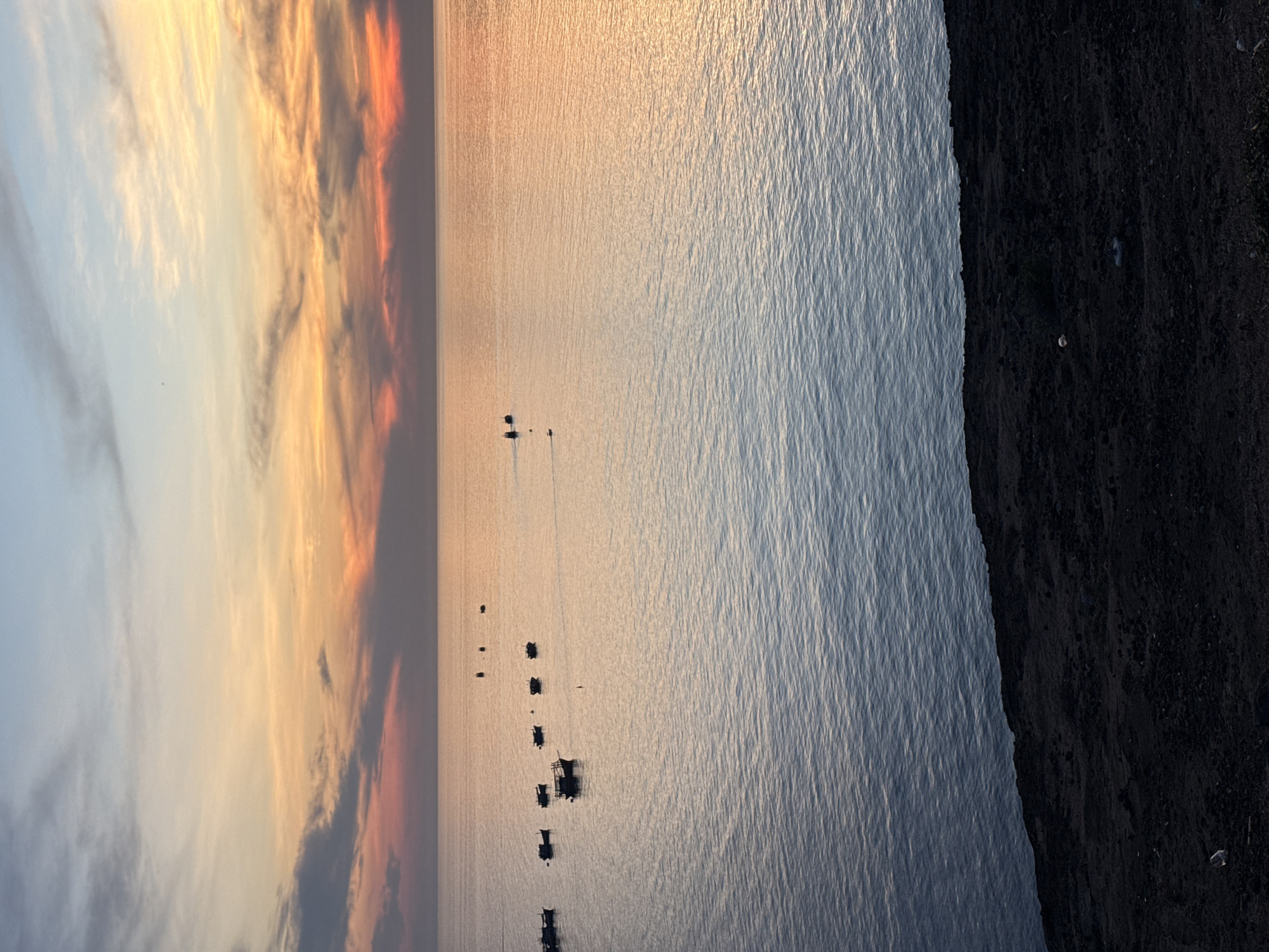 The image depicts a serene sunset scene at sea, with a cluster of small boats anchored in the calm water. The sky displays a gradient of colors, transitioning from a soft blue at the top to warm hues of orange and pink as it meets the horizon. The water reflects the colors of the sky, creating a tranquil and picturesque atmosphere. The foreground is dominated by a dark, rocky shoreline, adding a sense of depth and contrast to the composition. The overall impression is one of peacefulness and natural beauty. This scene is characteristic of the coastal areas of Lý Sơn Special Administrative Region, Vietnam, known for its stunning sunsets and fishing villages.