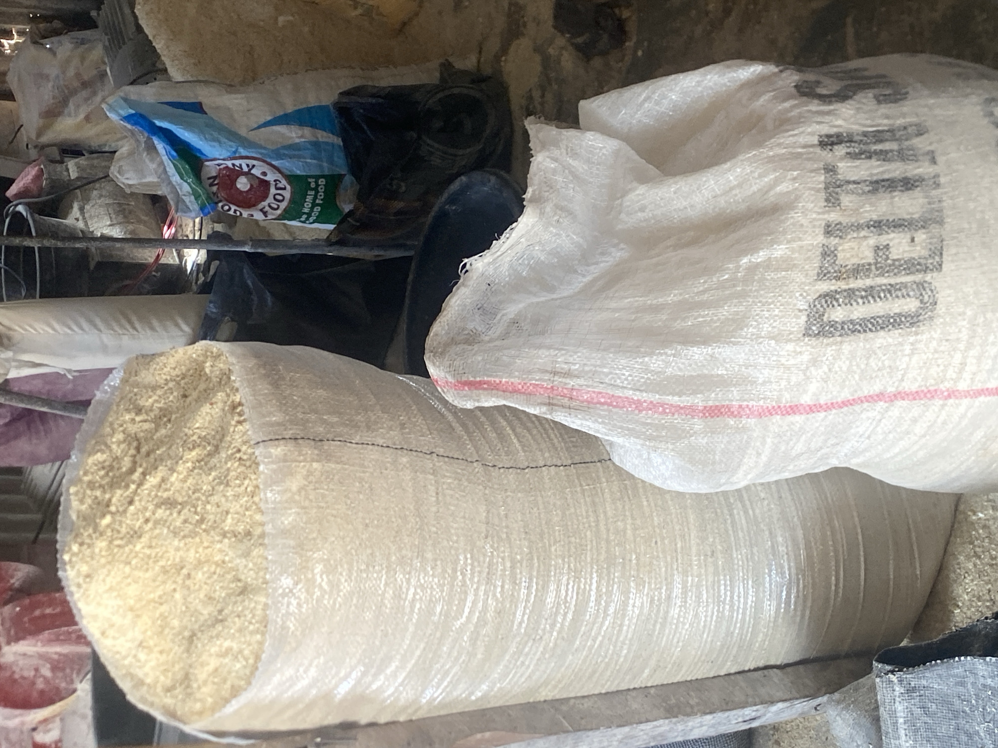 The image depicts a storage area in Bukuru, Nigeria, likely a warehouse or a market stall, filled with sacks of grain or other dry goods. A large, light-colored woven sack is prominently positioned on the left, filled to the brim with a pale yellow granular substance. To the right of this, a smaller, white woven sack is visible, bearing the black lettering "DELTA S" and red vertical stripes. Another sack with blue and white branding, featuring a circular logo with "GOOD FOOD" and the text "THE HOME OF GOOD FOOD," is also present, partially obscured. The background shows more sacks and storage containers, suggesting a commercial or agricultural storage setting. The floor appears to be concrete or dirt, with scattered debris, indicative of a working storage environment. No people are visible in the frame.