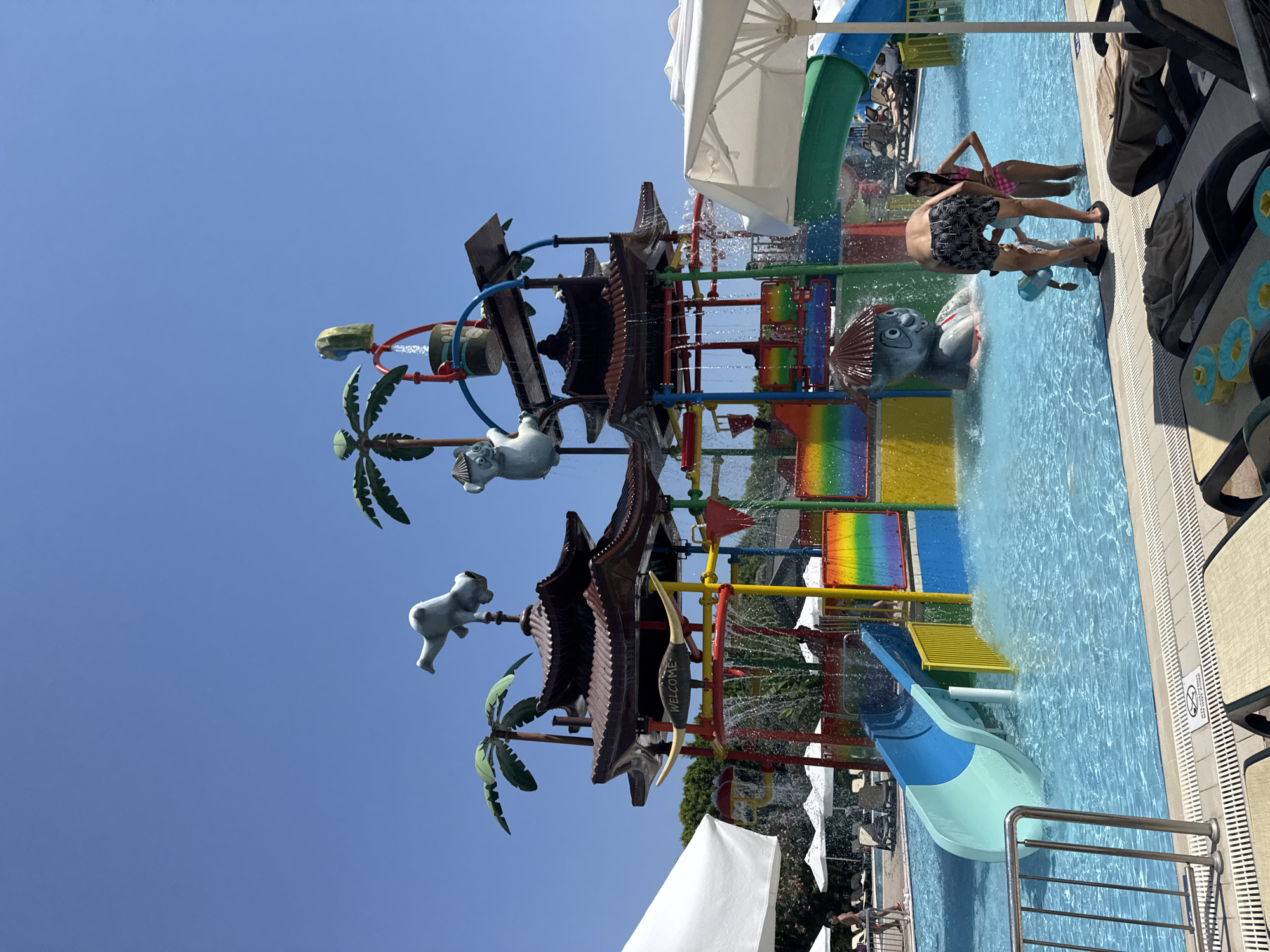 In Aksu, Turkey, a water park features a multi-level play structure with Asian-inspired roofs, colorful slides, and water features. Two children and an adult are in the shallow pool. The adult, wearing dark patterned swim shorts, faces away from the camera, bending down towards a younger child holding a blue water toy. A girl in a pink swimsuit stands nearby, facing the water structure. The pool area also shows lounge chairs and white umbrellas, with lush greenery in the background. The sky is clear blue.