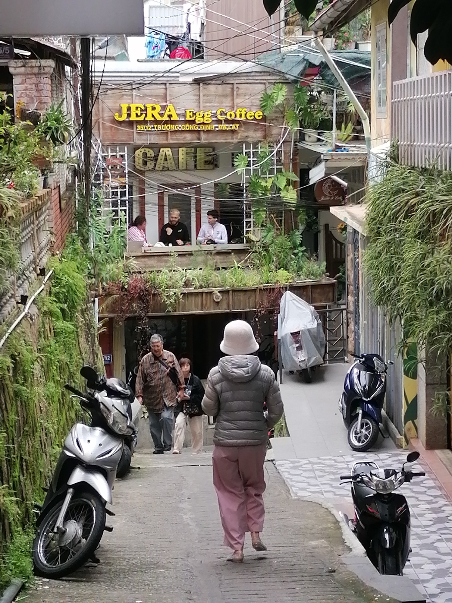 The image depicts a narrow, sloped street in Da Lat, Vietnam, with buildings on either side and several parked motorcycles. In the foreground, a person wearing a light-colored puffer jacket and pink pants walks away from the camera. To the left, a silver scooter is parked against a moss-covered wall. Further up the street, an older man in a plaid shirt and a younger person in dark clothing are walking. Above them, a sign for "JERA Egg Coffee" is visible, with "CAFE" written below it. People are seen sitting at tables inside the cafe on an upper level. The street is lined with plants and wires, suggesting a dense urban environment. Another person is walking towards the camera in the background. Several motorcycles are parked along the right side of the street, one of which is covered with a gray tarp. The architecture suggests older buildings with balconies and multiple stories.