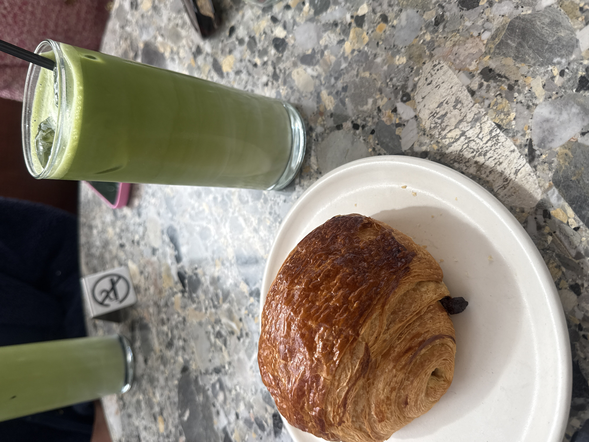The image depicts a table scene, likely in a cafe or restaurant. There is a drink of green liquid in a tall glass with ice and a straw; the color and texture suggest matcha latte, although this cannot be definitively confirmed. There is also a white plate that holds a pain au chocolat. The items rest on a marble-like countertop. The scene appears to be set in Los Angeles, United States of America. No people or interactions are distinctly visible, only portions of a person are visible at the bottom and left of the frame.