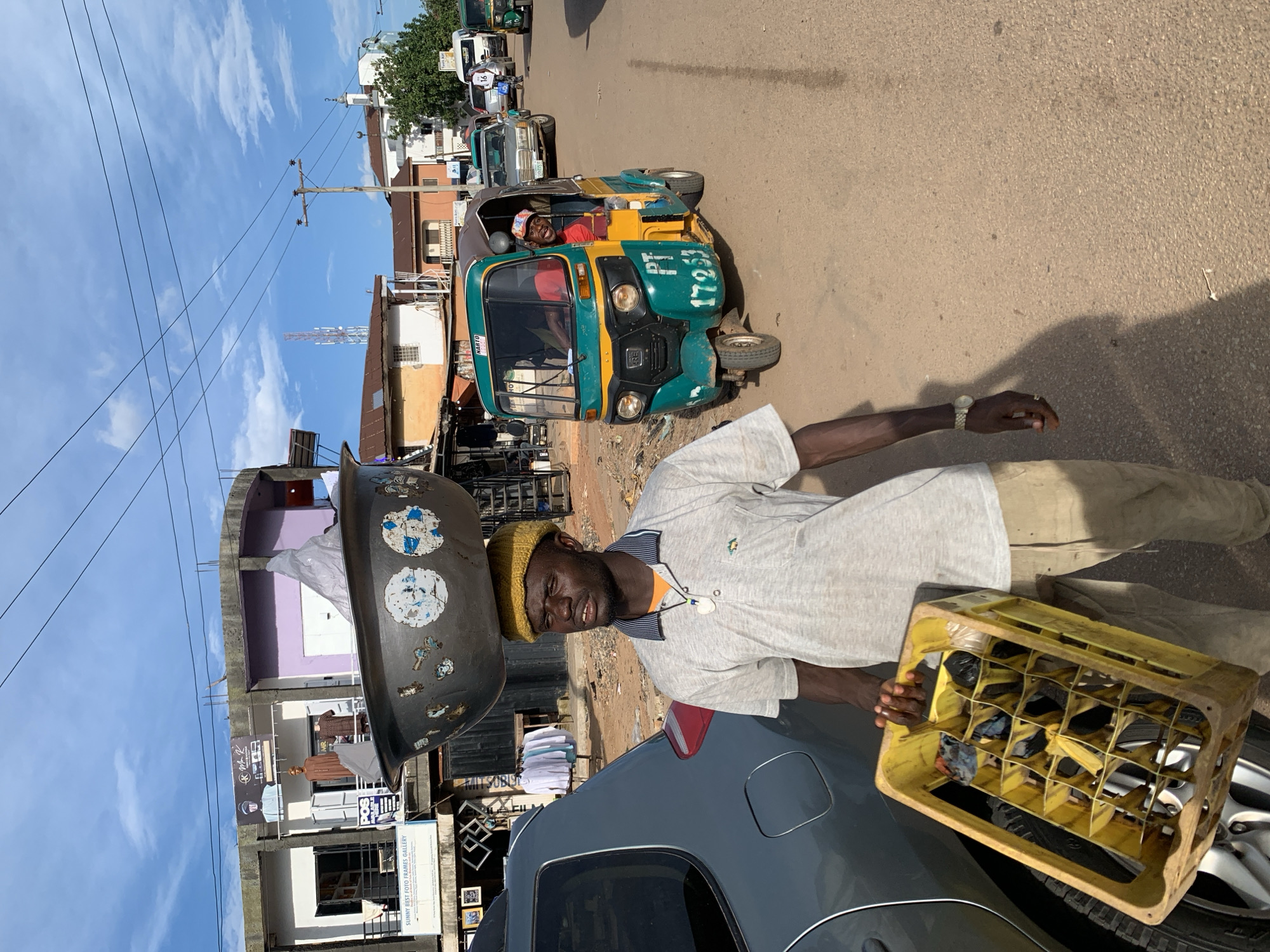 A man balances a large metal basin on his head while carrying a yellow plastic crate filled with bottles and other items. He wears a light gray polo shirt, tan pants, and a yellow knit cap. In the background, a green and yellow auto-rickshaw, also known as a Keke Napep, is driving down a dirt road. The driver, wearing a red shirt and a colorful cap, smiles at the camera. Several buildings, including shops and residences, line the street. The scene appears to be a marketplace or a busy street in Jos, Nigeria.