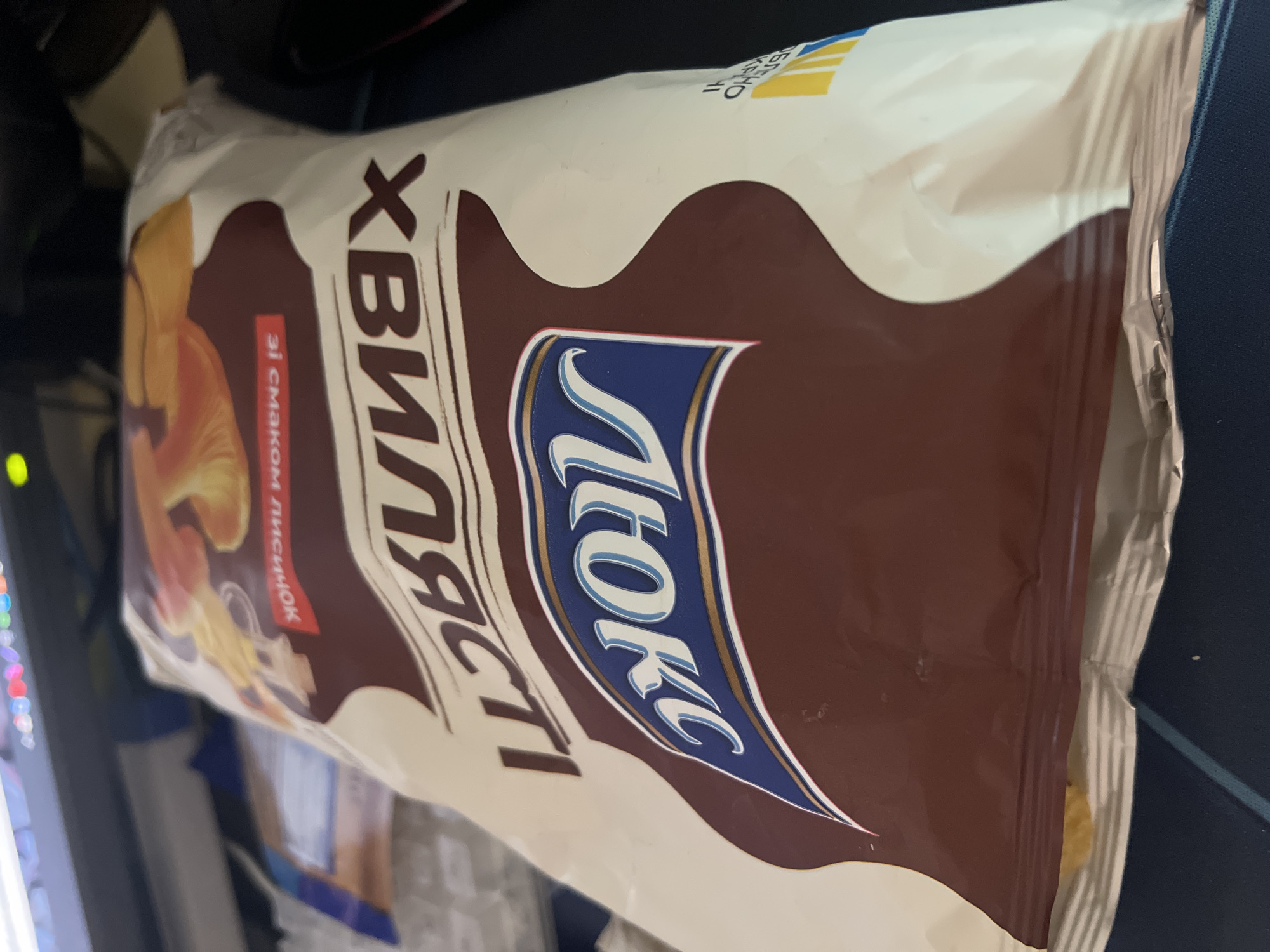 A bag of snacks, likely potato chips, is the subject of the image. The bag is primarily white and brown, with blue and gold accents. The brand name "ЛЮКС" is prominently displayed in large, stylized lettering on the front of the bag. Text in what appears to be Cyrillic script is visible on the bag, including "ХВИНАСТІ" and "ЗІ СМАКОМ ЛИСИЧОК". The bag is partially open at the bottom, revealing a few of the snacks inside. The bag is resting on a dark, textured surface, possibly a desk or table, with a blurry background that suggests an indoor environment.