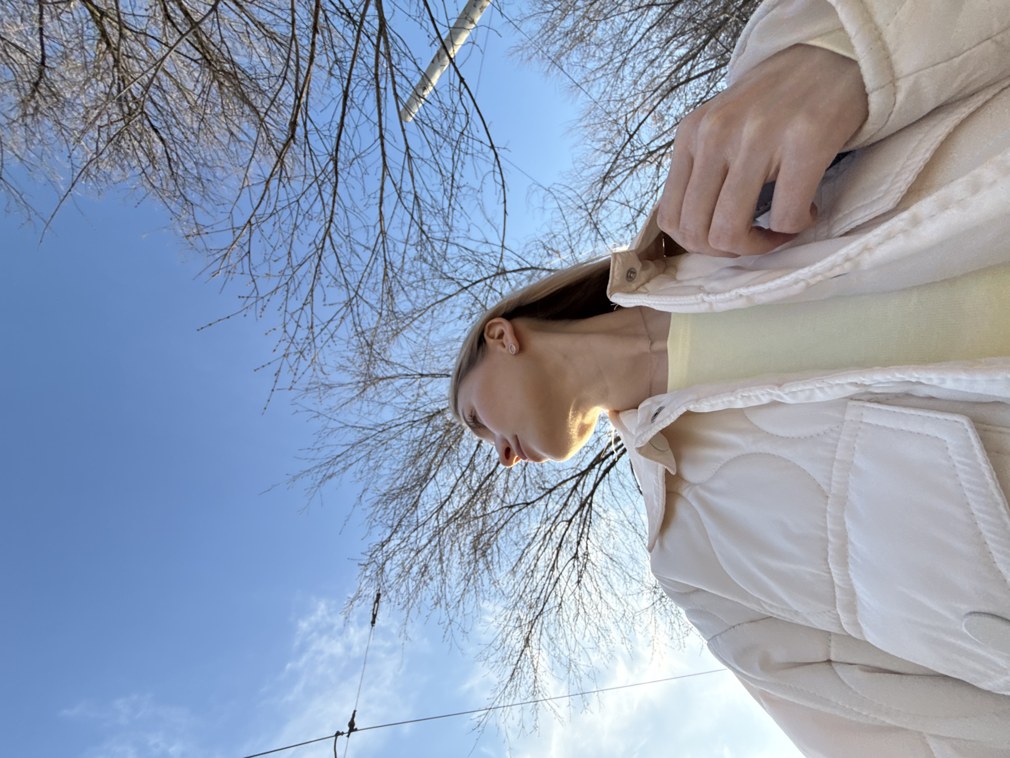 A low-angle shot captures a woman from the chest up, standing outside. She has fair skin, blonde hair pulled back, and wears a light yellow top under a cream-colored quilted jacket, which she partially holds closed with her right hand. Her face is in profile, looking downward. The background is dominated by a clear blue sky interspersed with bare tree branches. There are visible power lines in the lower portion of the frame.
There is insufficient information within the image to confirm a location or details about Izhevsk, Russia.