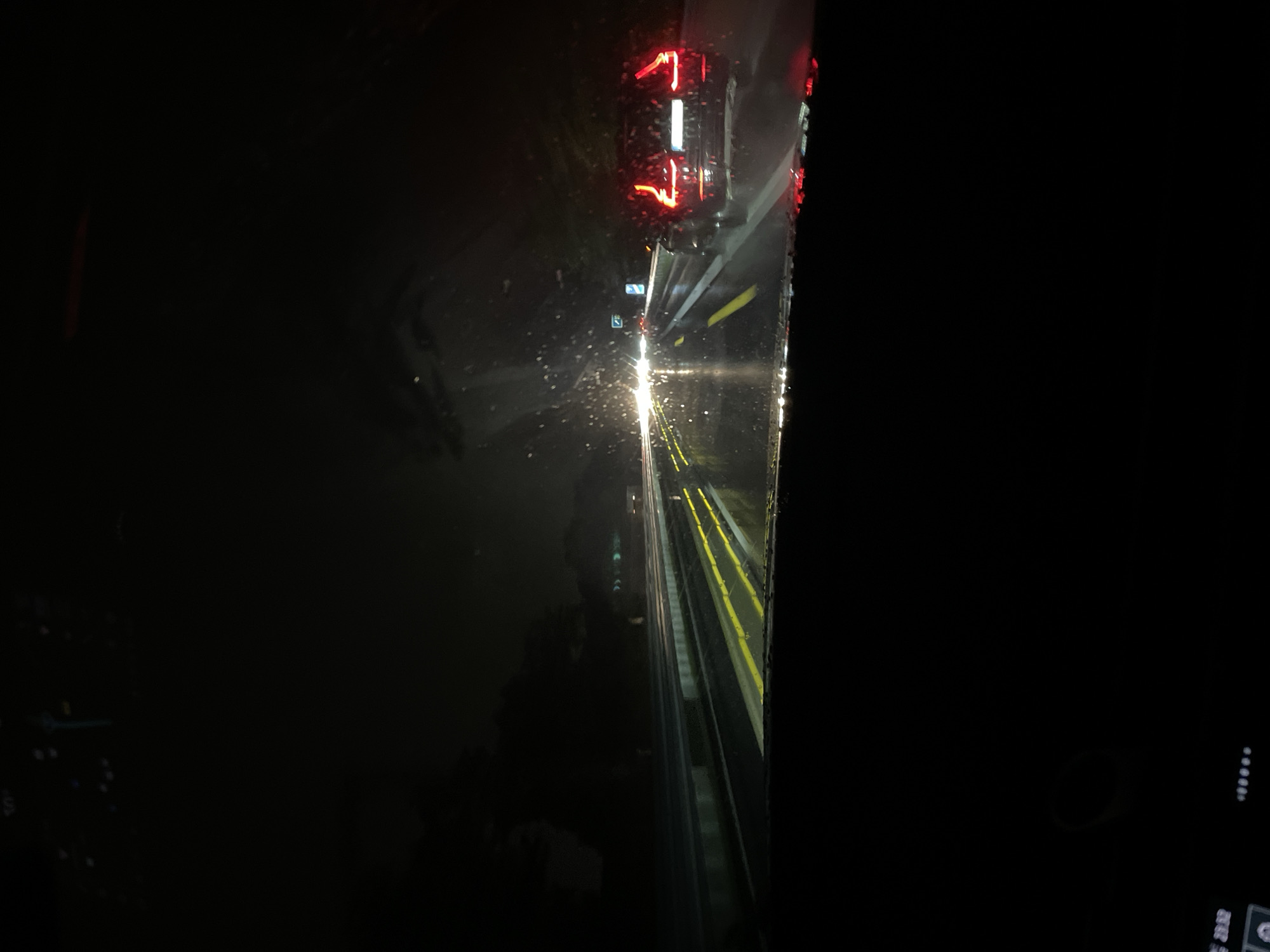 This image depicts a nighttime view from inside a vehicle on a wet road, possibly in or near Leverkusen, Germany. The road is illuminated by the vehicle's headlights and the taillights of a car ahead, a dark-colored SUV, which is partially visible on the right side of the frame. The road surface is reflective due to rain, with yellow lane markings visible. Distant lights and trees are visible on the left and right sides of the road, suggesting an urban or suburban environment. The windshield of the vehicle shows streaks of rain and light reflections. The overall atmosphere is dark and appears to be during a rainstorm.