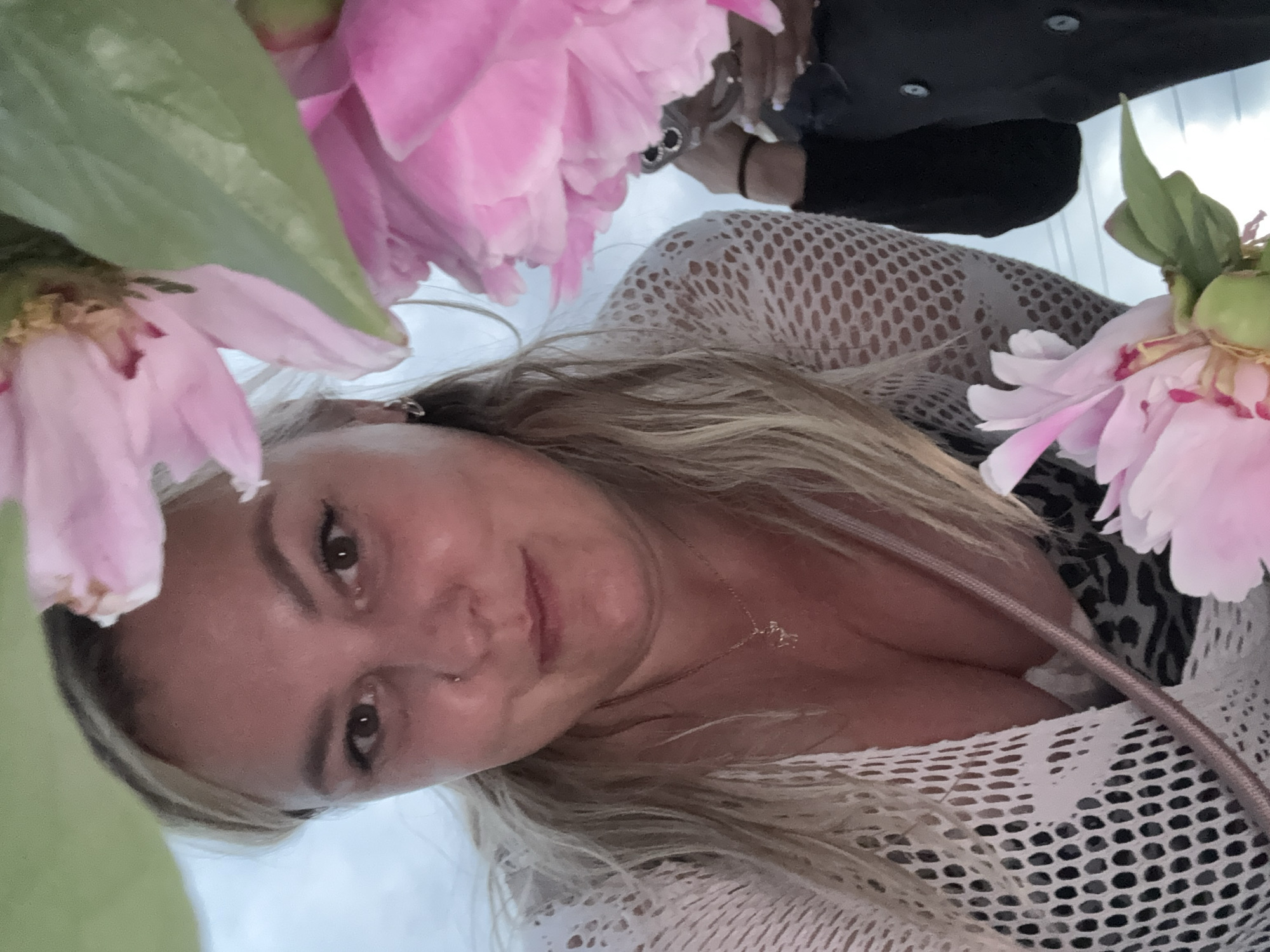 A woman with blonde hair and a nose ring is positioned in the foreground of the image, looking directly at the camera. She is wearing a white, open-knit top over a leopard-print garment. A delicate gold necklace with a small charm is visible around her neck. Partially obscuring her and the background are large pink flowers, possibly peonies, and green leaves. In the background, another person is partially visible, wearing a black jacket. The setting appears to be outdoors, with a bright sky in the distance, suggesting daytime. The image was taken in Chaikovsky, Russia.