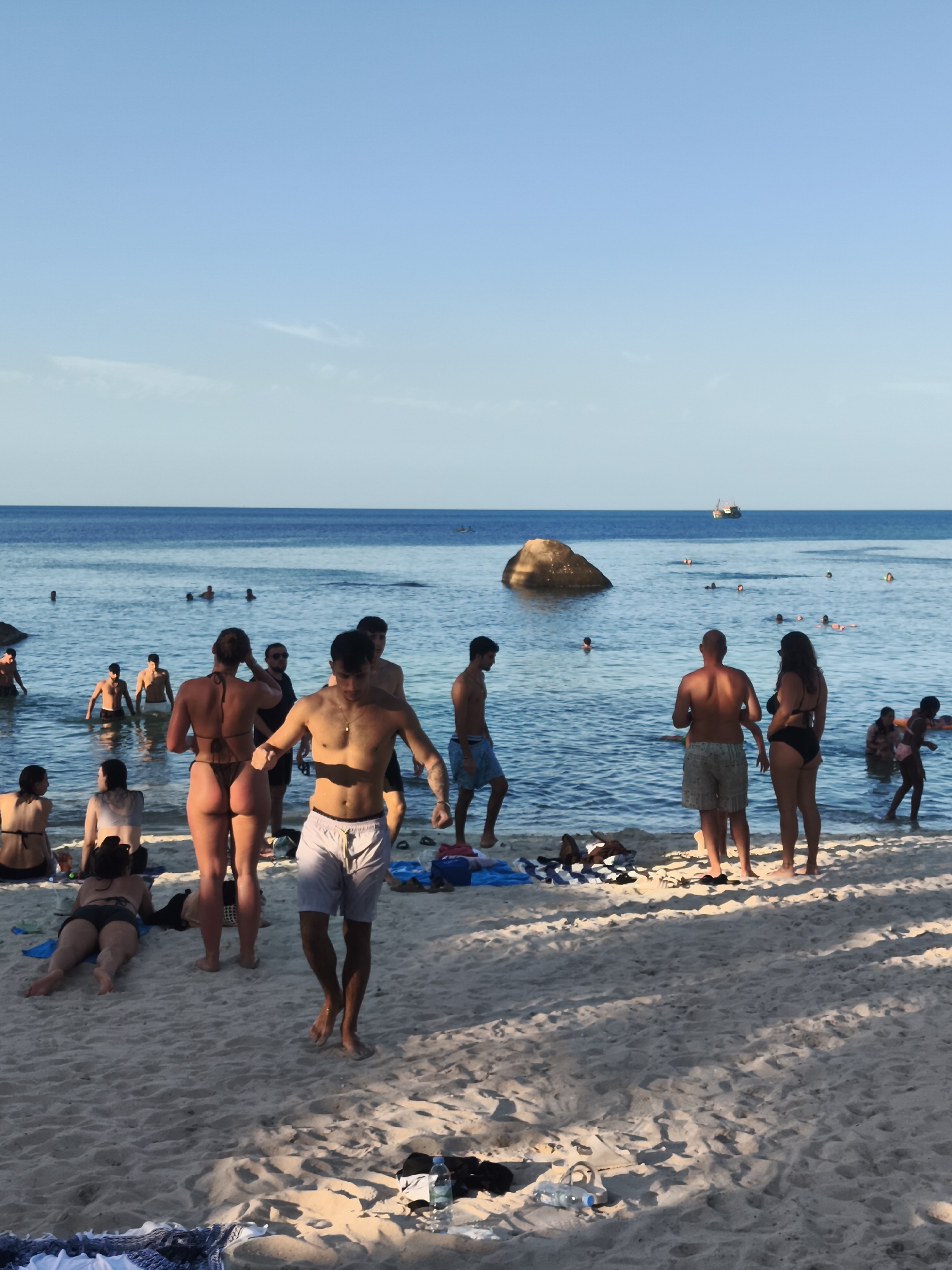 A photograph taken on a beach in Ko Samui District, Thailand, shows a group of people enjoying the ocean and sand. The sky is a clear blue. The water is calm and clear, with several people swimming and wading. On the beach, some individuals are sitting or lying on the sand, while others are standing and interacting. In the distance, a boat is visible on the water. The sand is light in color, and shadows are cast by people and objects, indicating it is likely late afternoon. The scene depicts a typical beach day with recreational activities.