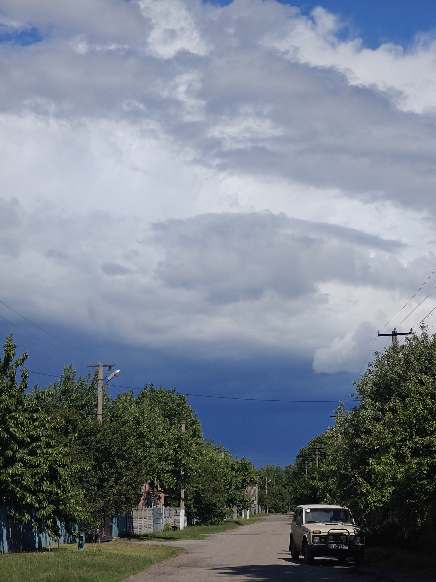The image depicts a street scene in Knyshivka, Ukraine. The road stretches into the distance, lined with trees on both sides. Power lines are strung along the street. A white Lada Niva car is parked on the right side of the road. The sky is filled with a mix of white and dark storm clouds, with a patch of dark blue sky visible in the distance, suggesting an approaching storm. A fence and some buildings are visible behind the trees on the left. The overall scene is rural or suburban.