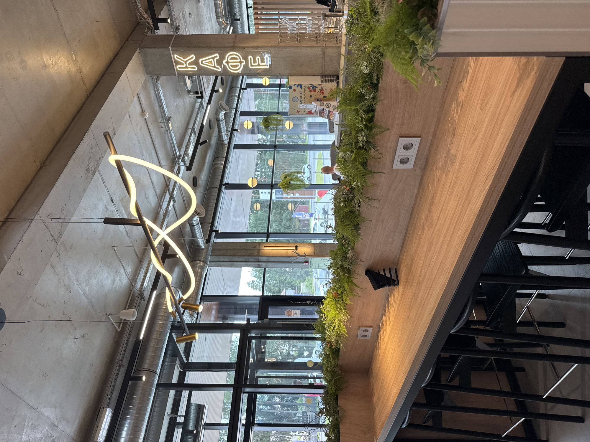 The image shows the interior of a modern cafe with an industrial aesthetic, featuring exposed concrete ceilings, metal ductwork, and large windows. A long, wooden counter runs along the foreground, with black bar stools positioned underneath. The counter is decorated with an abundance of lush green plants, creating a vibrant contrast with the raw materials. Electrical outlets are visible on the wooden wall behind the counter. In the background, through the windows, a street scene with cars and trees can be observed, indicating an urban or semi-urban environment. A neon sign spelling "КАФЕ" (Cafe) in Cyrillic letters is prominently displayed on a concrete pillar on the right side of the image. The overall ambiance suggests a contemporary and stylish cafe, possibly located in Lakhdenpokhya, Russia, given the Cyrillic signage. No individuals are clearly depicted or engaged in significant actions.