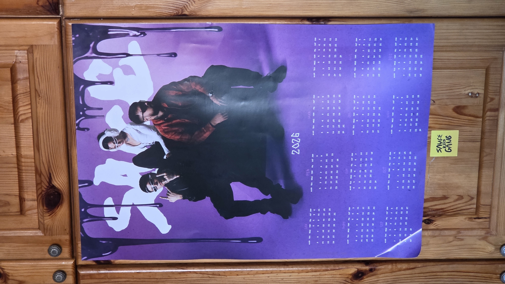 The image shows a 2026 calendar featuring the "Sauce Kids Gang," taped to a wooden cabinet. The calendar has a purple background with white abstract shapes and black "drips," and a picture of three individuals. A yellow sticky note with "SAUCE KIDS GANG" written in green ink is taped to the right side of the calendar.