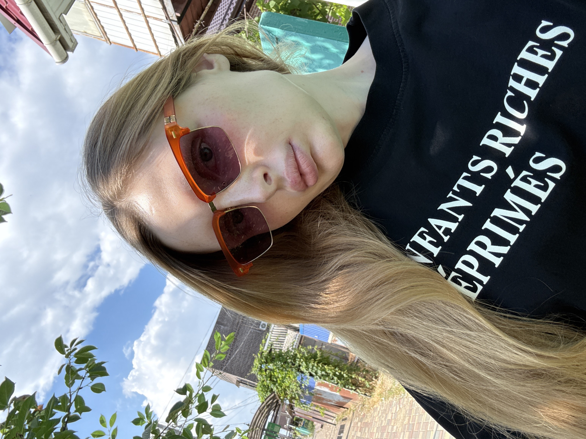 A young woman with long, blonde hair is taking a selfie outdoors on a sunny day. She is wearing sunglasses with orange frames and a black t-shirt with white lettering. The background shows trees, a building, and a cloudy blue sky.