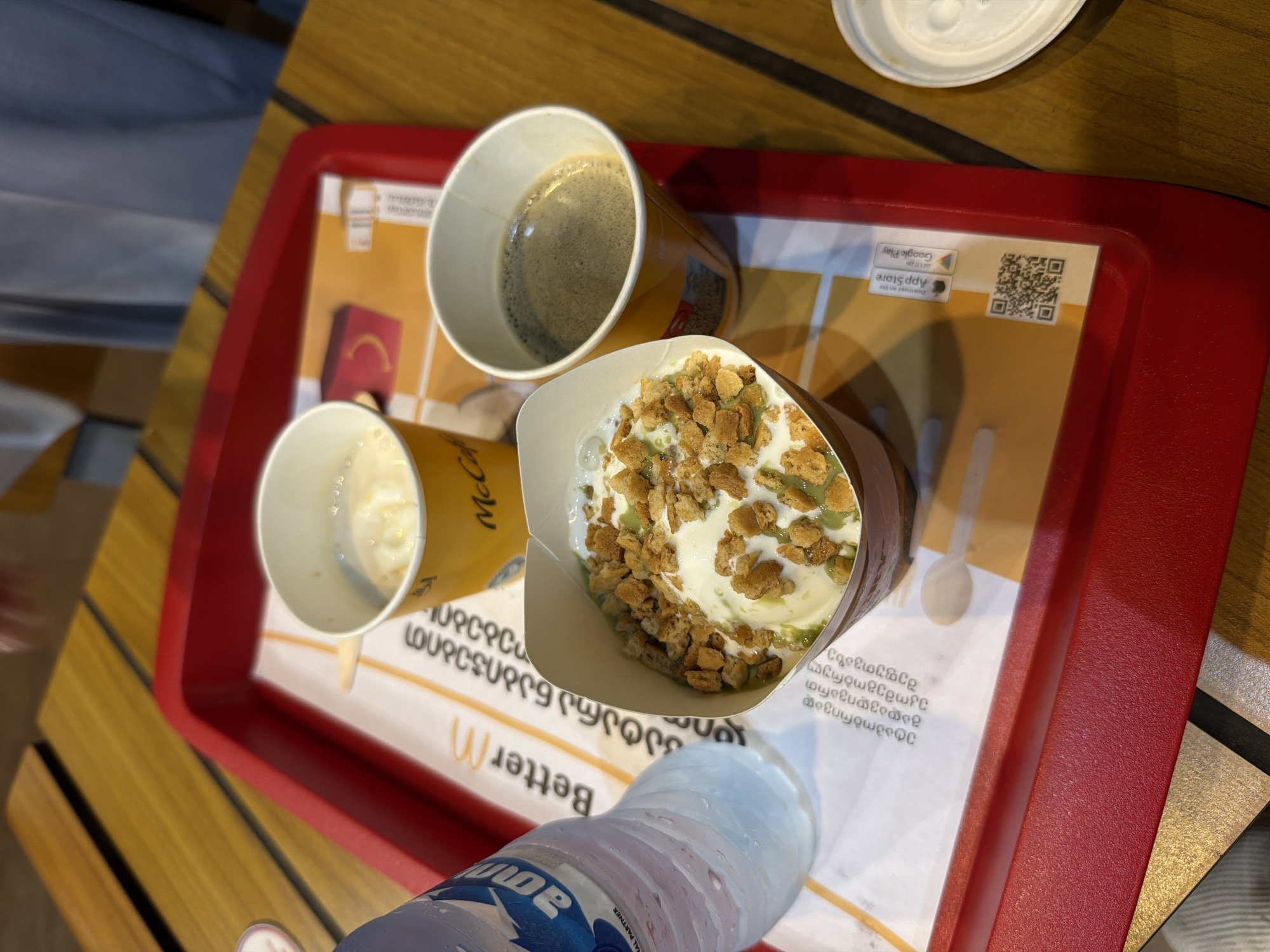 On a red tray, there are two yellow McDonald's cups, one with ice cream topped with crumbles, and another with coffee. Next to them is a partially visible bottle of Aquafina water. The tray is on a wooden table, and the McDonald's branding is visible on the paper liner underneath the cups. The scene is likely inside a McDonald's restaurant in Batumi, Georgia, given the Georgian text on the paper liner.