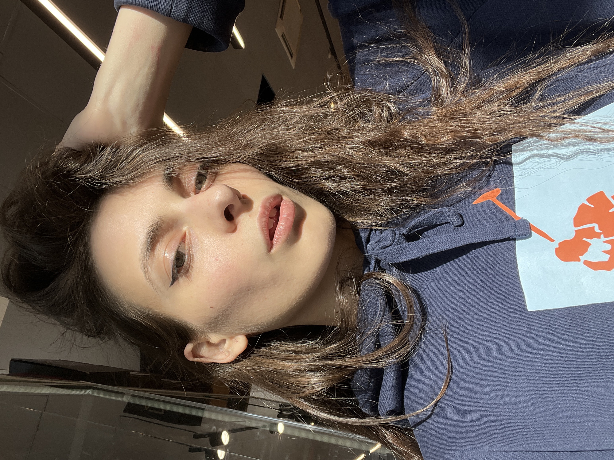 A young woman with long brown hair lies in a supine position, facing upwards at an angle. She is wearing a dark blue hooded sweatshirt with a light blue and orange design on the chest. Her left hand is raised above her head, touching her hair. She has fair skin, and her face is well-lit. Her lips are slightly parted, and she has eyeliner on her upper eyelids. The background is partially visible, showing a reflective surface that could be glass or polished material, with indistinct shapes and light sources. The overall environment suggests an indoor location. The location in the city of Krasnoyarsk, Russia is not discernable from the image.