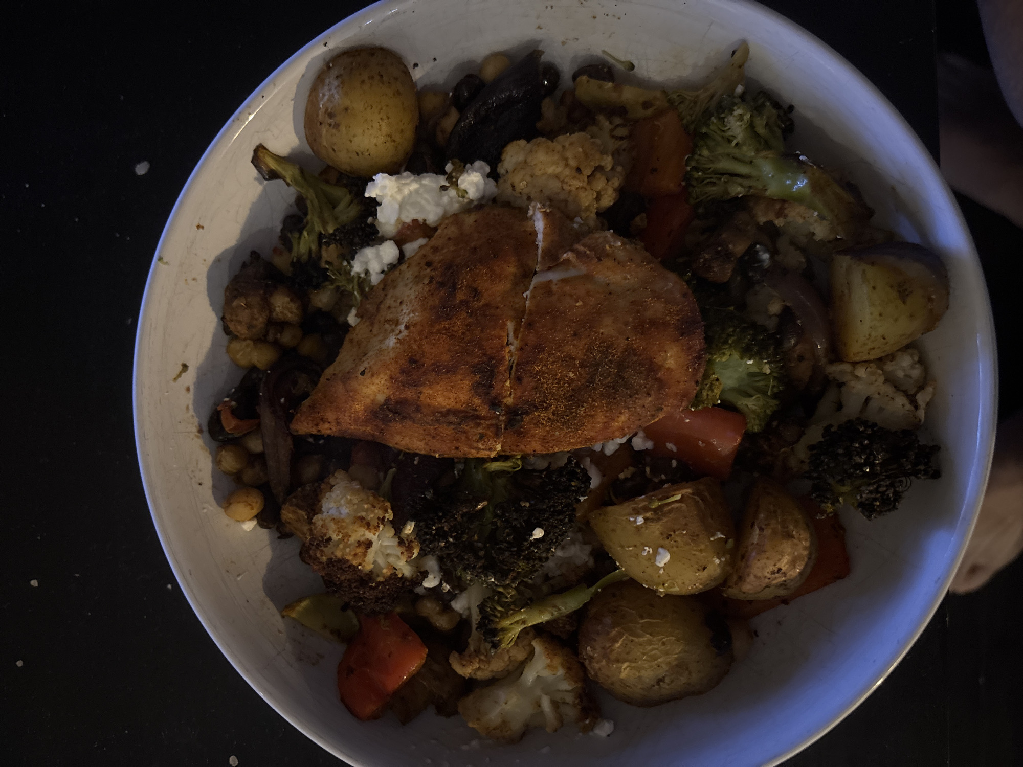A top-down view of a white bowl filled with food. The bowl contains a seasoned chicken breast, roasted vegetables including potatoes, cauliflower, broccoli, and red peppers. There are also chickpeas, olives, and crumbled feta cheese. The bowl is placed on a black surface.