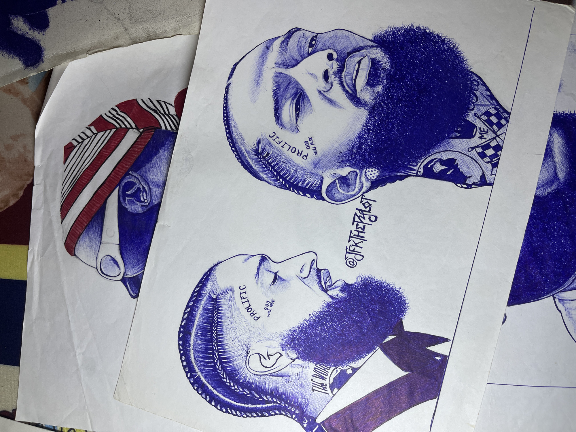 The image shows three ballpoint pen drawings on white paper. Two drawings depict the same man, likely the deceased rapper Nipsey Hussle, in profile and facing forward, with the word "PROLIFIC" tattooed on his face and stylized braids. The drawings are rendered in shades of blue ink. The third drawing shows a man wearing a red, white, and black head wrap, rendered in blue ink. The drawings are stacked on top of each other and other papers. The location, Jos, Nigeria, cannot be determined from the visual information.