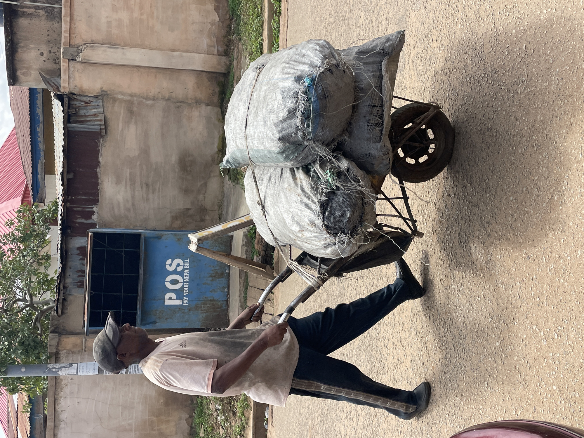 The image captures a man pushing a two-wheeled handcart heavily loaded with large, grey, woven bags along a coarse-grained, paved path. The man, viewed from his left side, is dressed in a light-colored t-shirt featuring an "Adidas" logo, dark trousers with side stripes, and a grey cap. He leans into the task, gripping the cart's handles. The bags, likely containing bulk materials such as charcoal, are tied down with various ropes and appear substantial.
The setting is an outdoor urban or semi-urban environment in Jos, Nigeria, characterized by bright daylight. In the background, aged concrete buildings with visible wear and tear, and corrugated metal roofs in shades of red and blue, line the street. A significant detail is a blue utility box or kiosk positioned behind the man, emblazoned with the white text "POS" and "PAY YOUR NEPA BILL," strongly rooting the scene in Nigeria. Green trees and shrubs are also visible to the left. The scene portrays an everyday activity of manual transport.