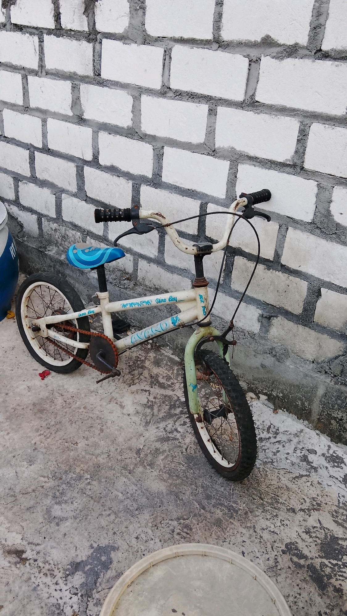 A rusted, cream-colored children's BMX bicycle is positioned in front of a white brick wall, with its tires resting on a concrete surface. The bicycle has a blue seat with a wavy pattern and green handlebars. The chain and pedals are rusty, indicating age and disuse. The front fork of the bicycle is painted green. The wall behind the bicycle is constructed from rectangular white bricks with gray mortar. The concrete ground is stained and weathered. A large, light-colored circular object, possibly a bucket lid, is visible in the foreground. A blue cylindrical object is partially visible to the left of the bicycle. The scene is outdoors, likely in a yard or alleyway in Banjarsari, Indonesia, given the brickwork and the type of bicycle, common in many Indonesian urban environments.