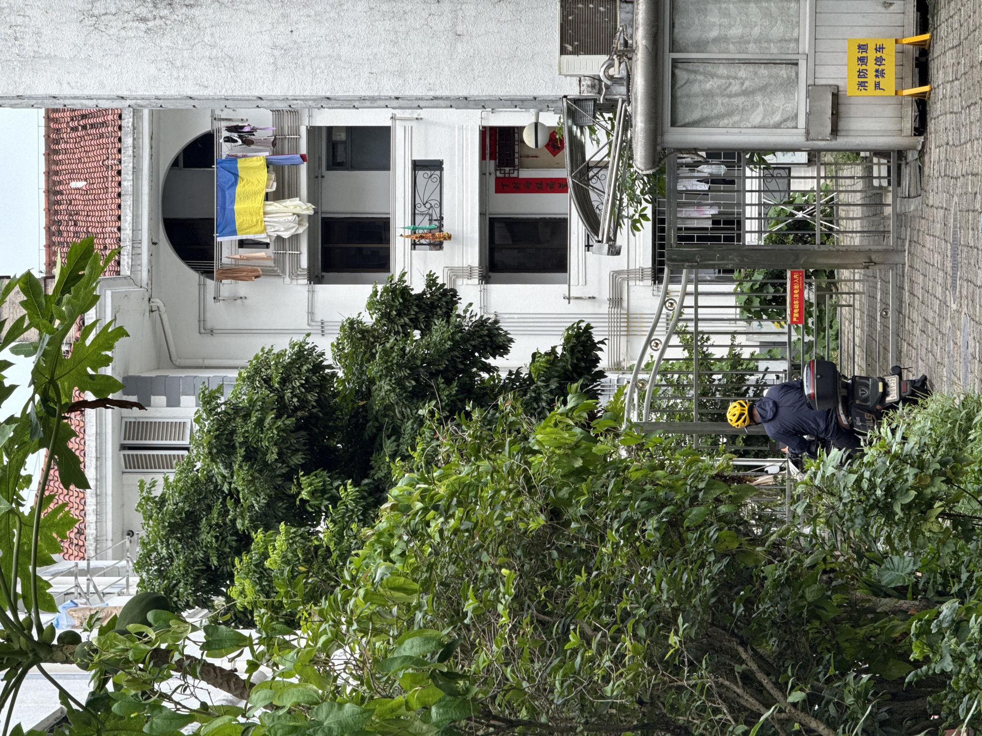 A person wearing a yellow helmet and a dark blue jacket rides a scooter towards a gated entrance of a white apartment building, which has a red-tiled roof and a balcony displaying a Ukrainian flag and laundry. Green foliage partially obscures the building and the scooter rider. A small security booth with a yellow sign bearing Chinese characters is visible to the right of the gate. The ground is paved with cobblestones.