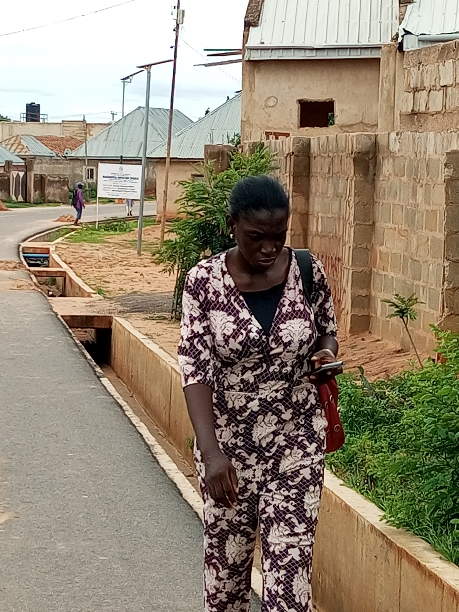 The image depicts a woman walking along a paved path next to a concrete drainage channel. She is wearing a patterned outfit and carrying a red bag. In the background, there are buildings with corrugated metal roofs and a stone wall. A signpost with text in English is visible further down the path, along with another person walking in the distance. The overall setting suggests an urban environment in Gombe, Nigeria, with a mix of residential and possibly institutional buildings. The sky is overcast, indicating a cloudy day.