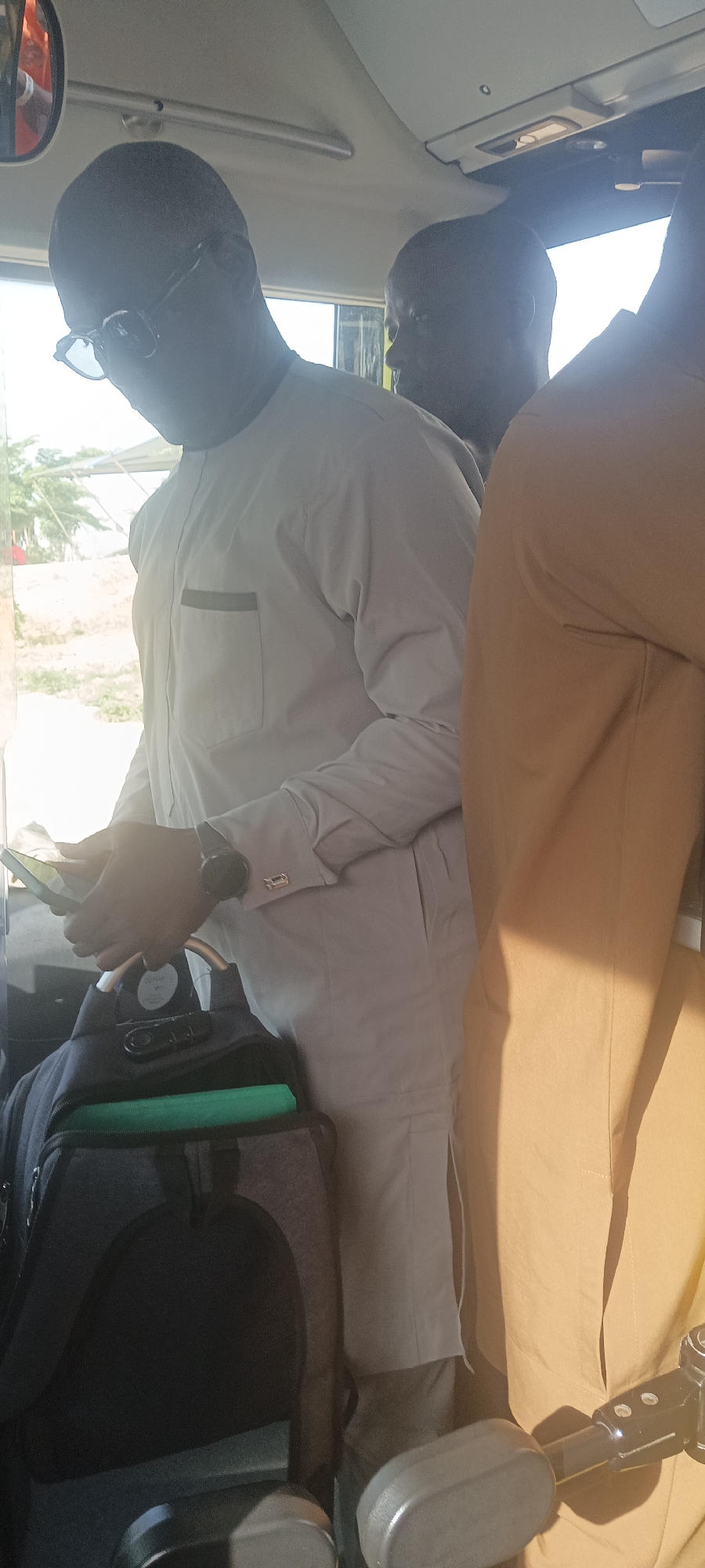The image shows three dark-skinned men inside a vehicle, possibly a bus, in Jos, Nigeria. The man in the foreground is wearing glasses and a light-colored, long-sleeved shirt and matching pants. He is looking down and appears to be using a smartphone. He has a wristwatch on his left wrist, and cufflinks visible on his sleeves. A black and grey backpack with a green item on top of it is in front of him. Partially visible behind him is another dark-skinned man in a brown shirt. The interior of the bus is visible, including a window and ceiling elements. Outside the window, there is a glimpse of greenery and a building in the background.
