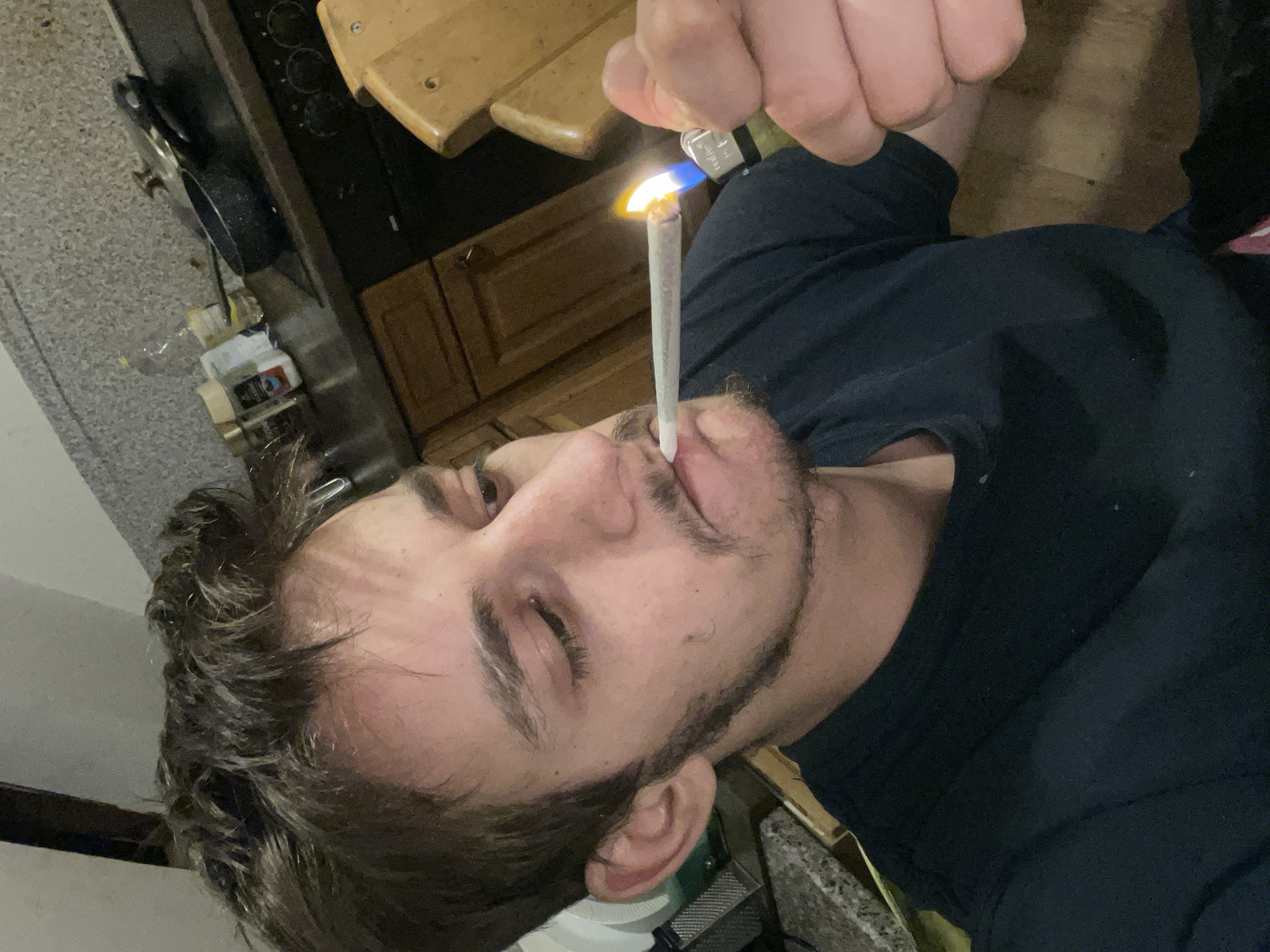 A male with light brown hair and a beard is lighting a cylindrical white object with a blue and yellow flame from a lighter held in his right hand. The scene appears to be indoors, possibly in a kitchen based on the wooden cabinets, countertops, stovetop, and cooking equipment visible in the background. The location is Unknown, Unknown. The male is wearing a dark-colored shirt. The object being lit appears to be held in his mouth.