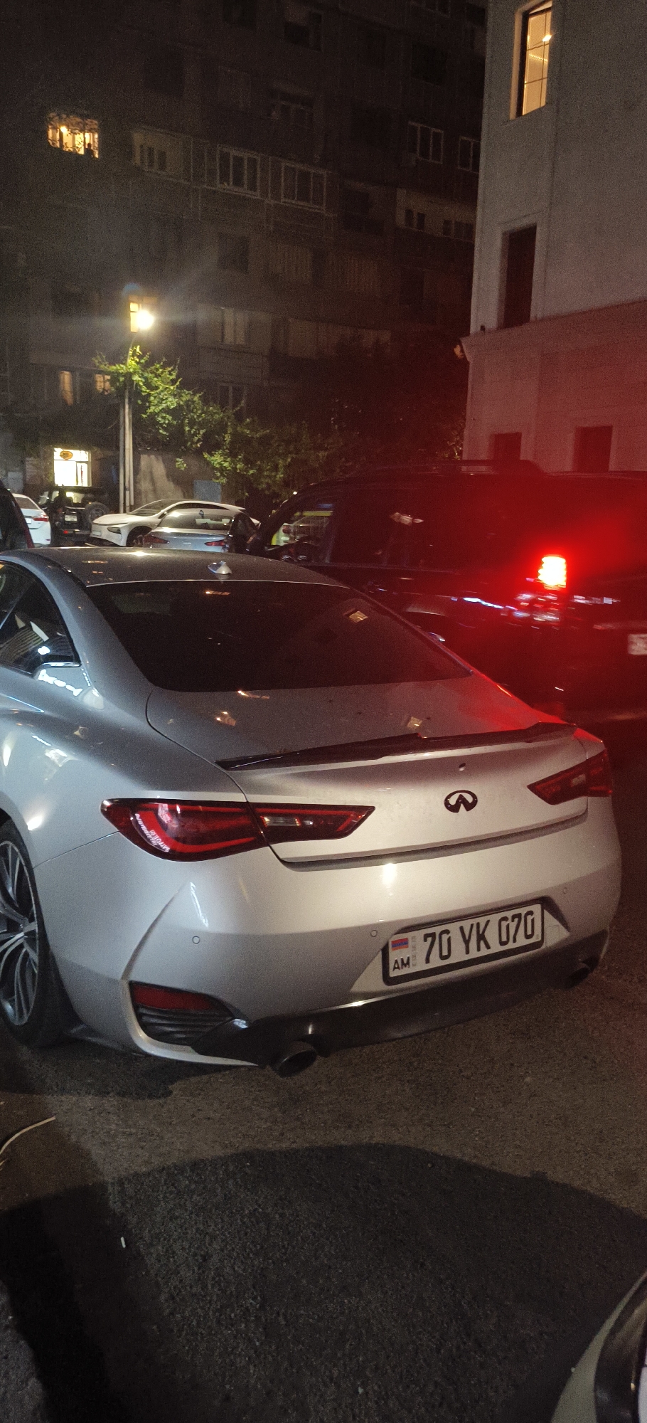 In an urban setting at night, a silver Infiniti Q60 coupe is parked, facing away from the camera. The car has a spoiler on the trunk and a license plate reading "70 YK 070" with Armenian markings. To the right of the Infiniti, a black SUV with a flashing red and white light on its roof is visible. In the background, a multi-story apartment building is illuminated with some windows lit. A streetlamp with a green tree nearby is also present. Other cars are parked in the vicinity, partially obscured.