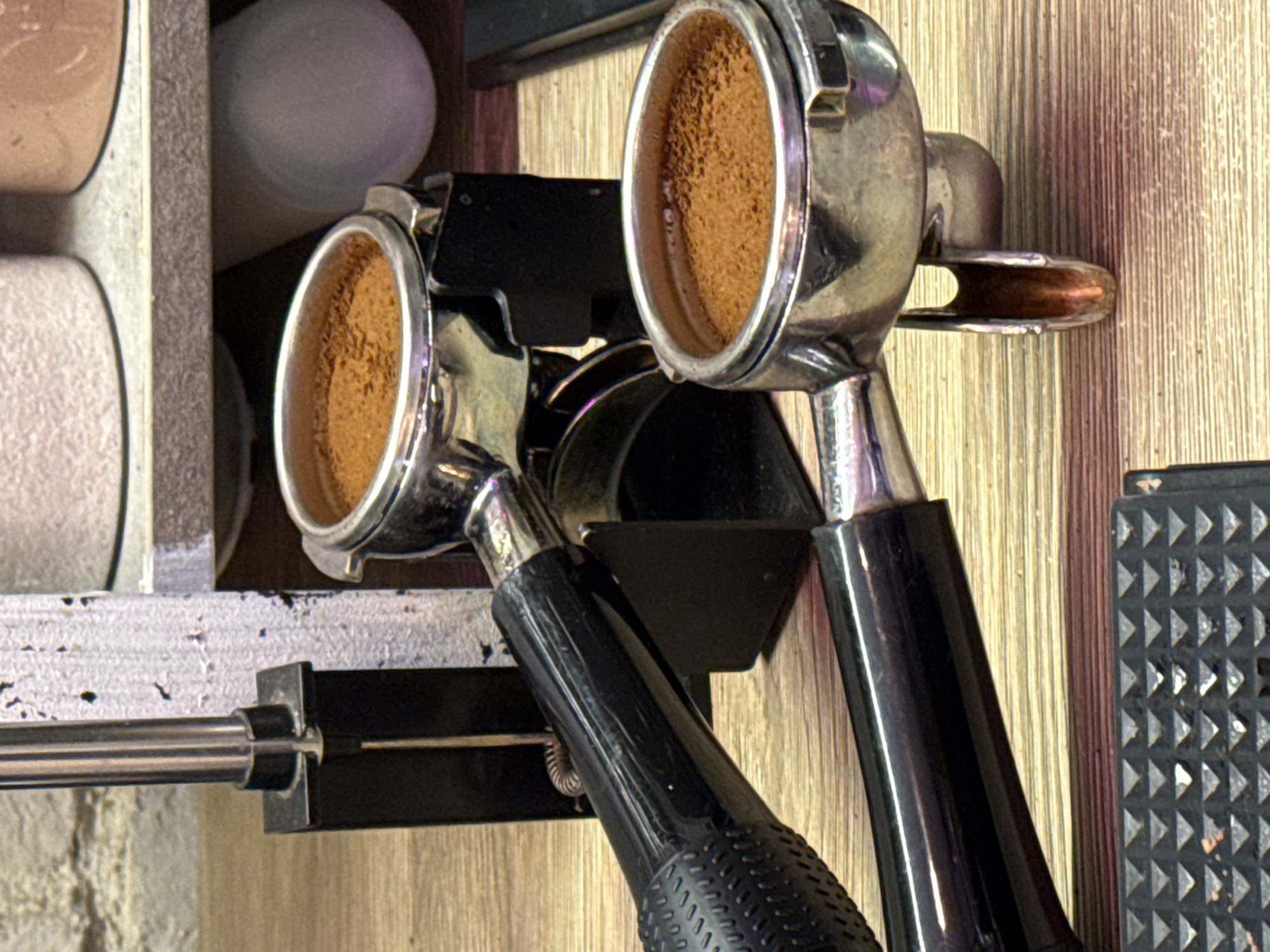 This is a close-up, eye-level shot of two espresso portafilters filled with coffee grounds. The portafilters are made of shiny metal with black handles. One portafilter is positioned in front of and slightly below the other, and both are resting on a wooden surface. In the background, a vertical metal rod is visible, attached to a black base, and beyond that, a shelf with two containers that appear to be holding powders or granules. The overall scene suggests a coffee preparation area. There is no information to place this image in Moscow, Russia.