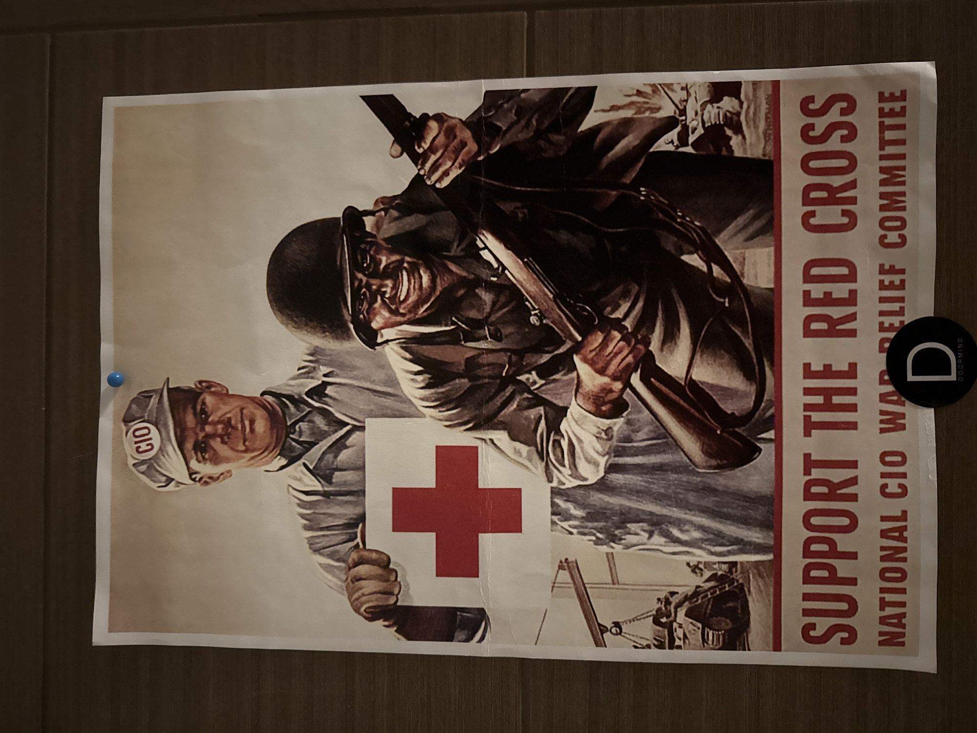 The image shows a poster with a wartime theme. The poster features two men: one is a worker with a cap labeled "CIO", and the other is a soldier holding a rifle. The worker is holding a large Red Cross symbol. The background shows machinery and other military figures. The poster text reads "SUPPORT THE RED CROSS" and "NATIONAL CIO WAR RELIEF COMMITTEE". The poster appears to be pinned to a wall, with a blue pin visible in the upper left corner and a round black object in the upper right corner. The location is unknown.