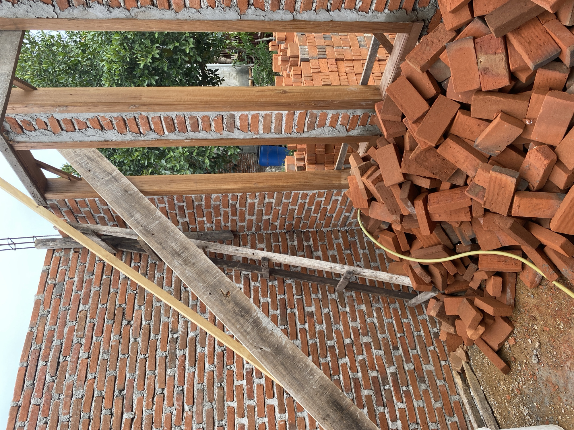 The image depicts the construction of a brick building in Banda Aceh, Indonesia. The scene is characterized by unfinished brick walls, wooden structural supports, and a pile of loose bricks in the foreground. A yellow hose snakes across the ground near the pile of bricks. In the background, stacks of bricks are visible, as are green trees and a blue barrel. The overall impression is one of ongoing construction work.