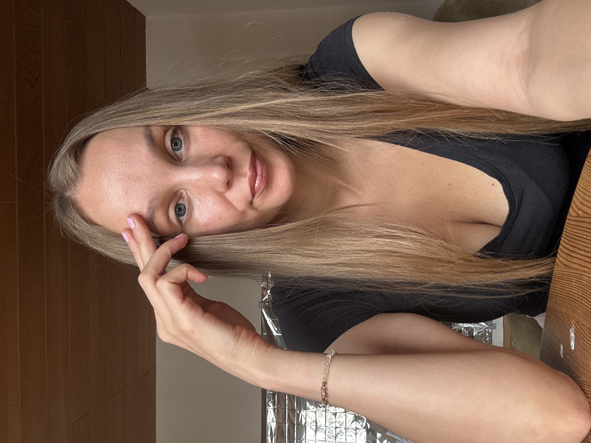 A blonde woman is seen from the chest up, sitting at a wooden table. She is wearing a black t-shirt and has a bracelet on her left wrist. Her right hand is resting on her forehead. The background shows a wooden ceiling and a wall with metallic foil. The setting appears to be an indoor environment, possibly a room in Überherrn, Germany.
