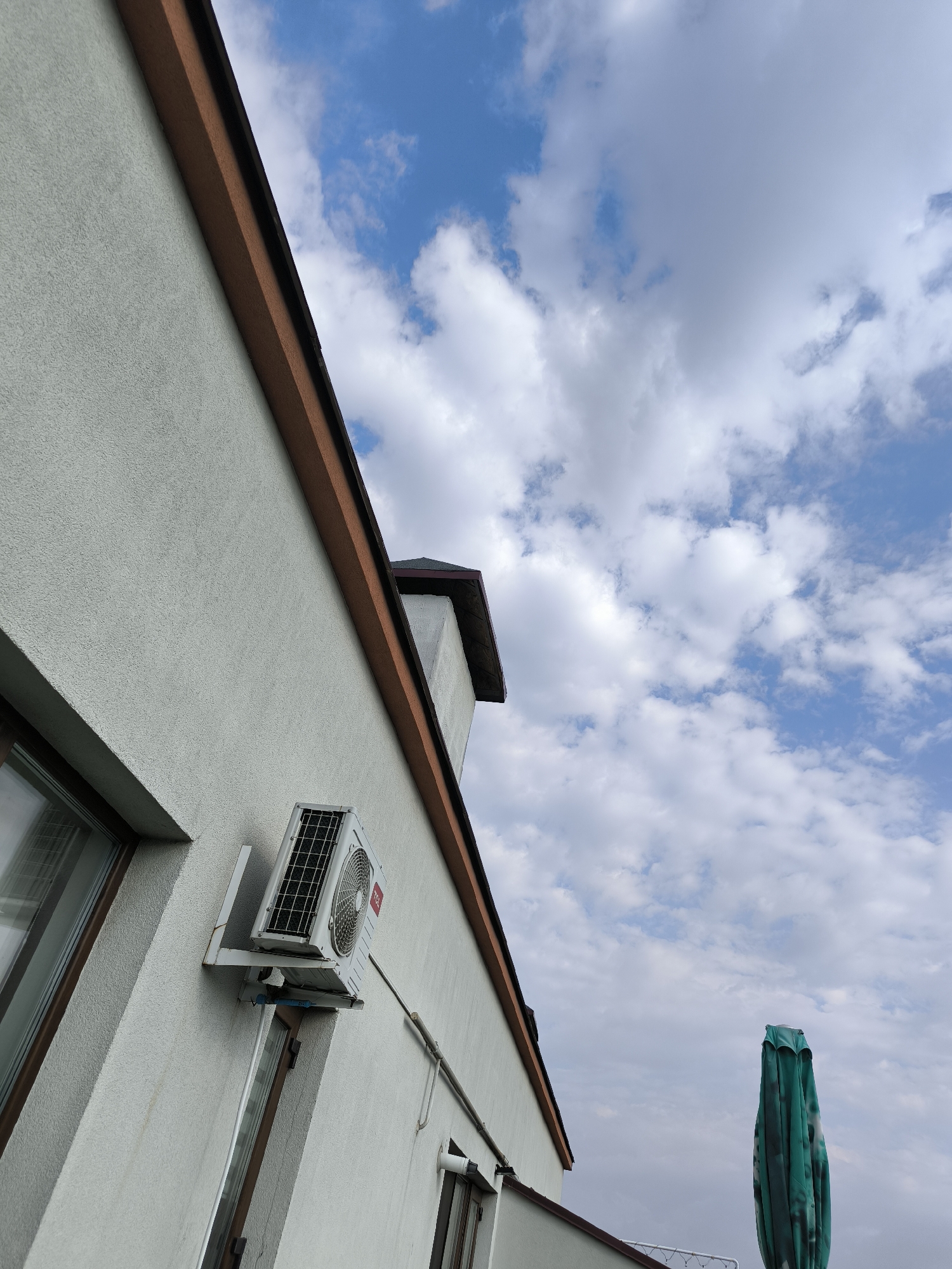 The image is a low-angle outdoor shot of the exterior of a building against a cloudy blue sky. On the textured, light-colored wall of the building, an outdoor unit of an air conditioner is mounted. A window is visible to the left of the air conditioner unit. The roofline of the building, with a brown fascia and a dark overhang, is visible along the top edge. A chimney with a dark cap extends upwards from the roof in the upper middle of the frame. To the right of the building, a closed, green patio umbrella stands vertically. The scene appears to be in a residential area, possibly a private house or apartment building. There are no people or animals present in the image. The lighting suggests it is daytime, with the sun partially obscured by clouds. The overall atmosphere is calm and ordinary. There is no specific information in the image to suggest it is located in Chișinău, Moldova.