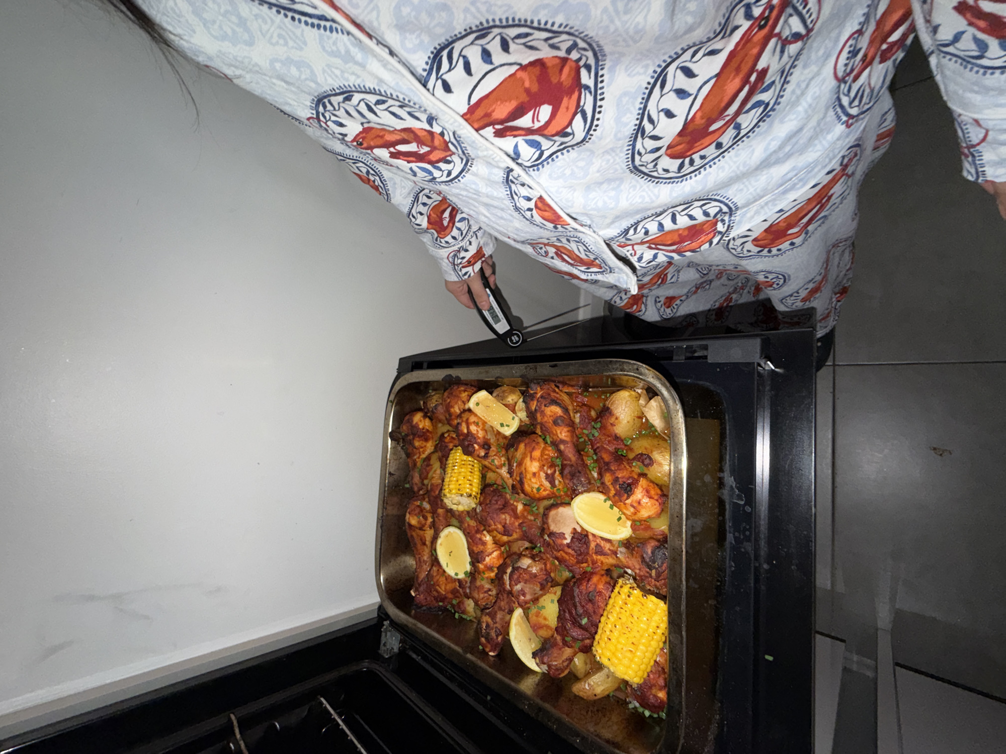 A person wearing pajamas with a lobster pattern is holding a digital thermometer, with the probe inserted into a large roasting pan filled with chicken pieces, potatoes, and corn on the cob. The roasting pan is sitting on the open door of an oven, which appears to be in a domestic kitchen. The background shows a white wall with some smudges.