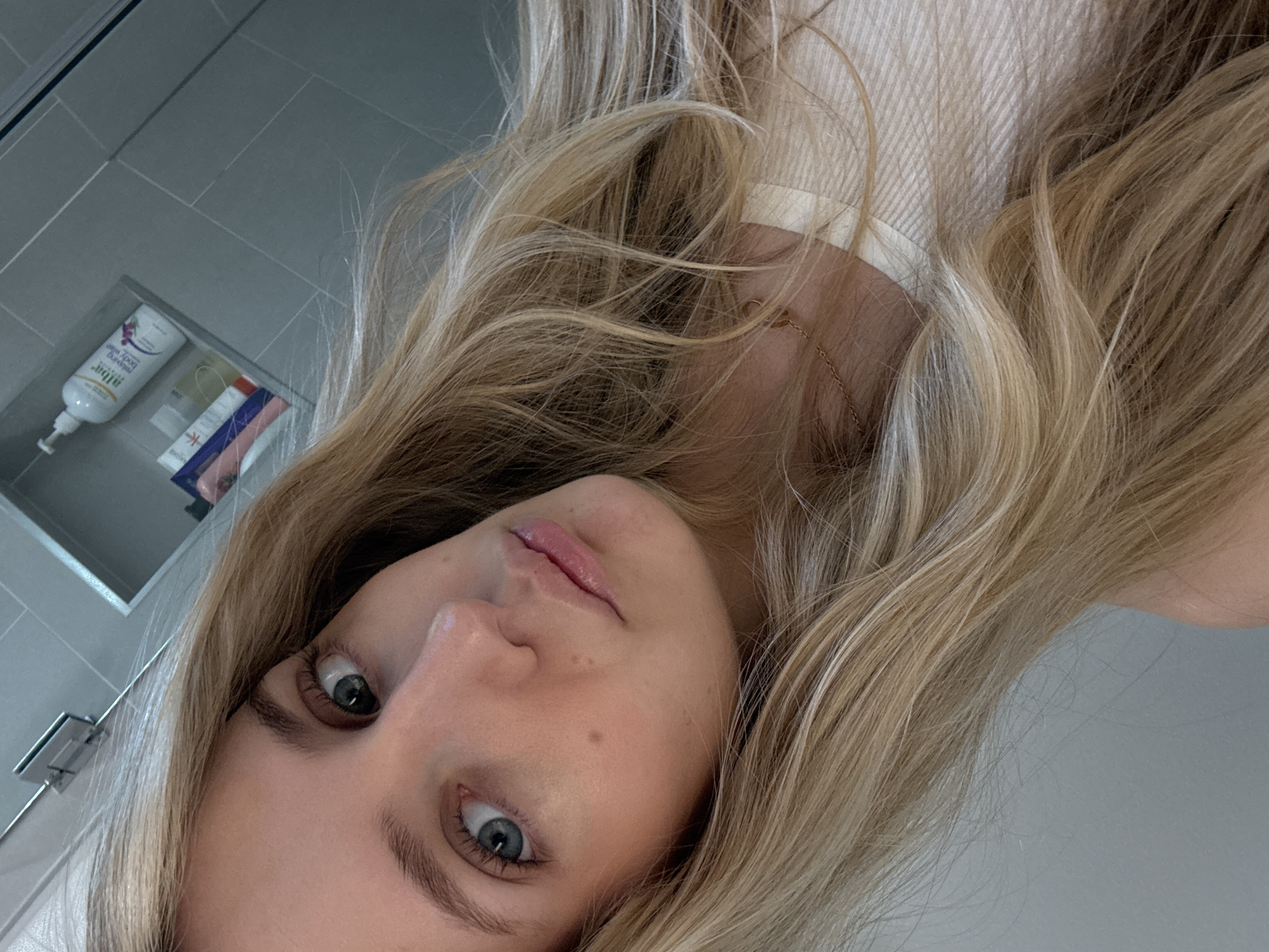 A young Caucasian woman with blonde hair and blue eyes is captured in a close-up selfie. She is wearing a light-colored top. The background appears to be a bathroom with tiled walls and a shelf containing various toiletries, including a bottle of "Alba Botanica Hawaiian Body Wash." The scene is lit with artificial light, creating a soft glow on her face. The image was likely taken indoors.