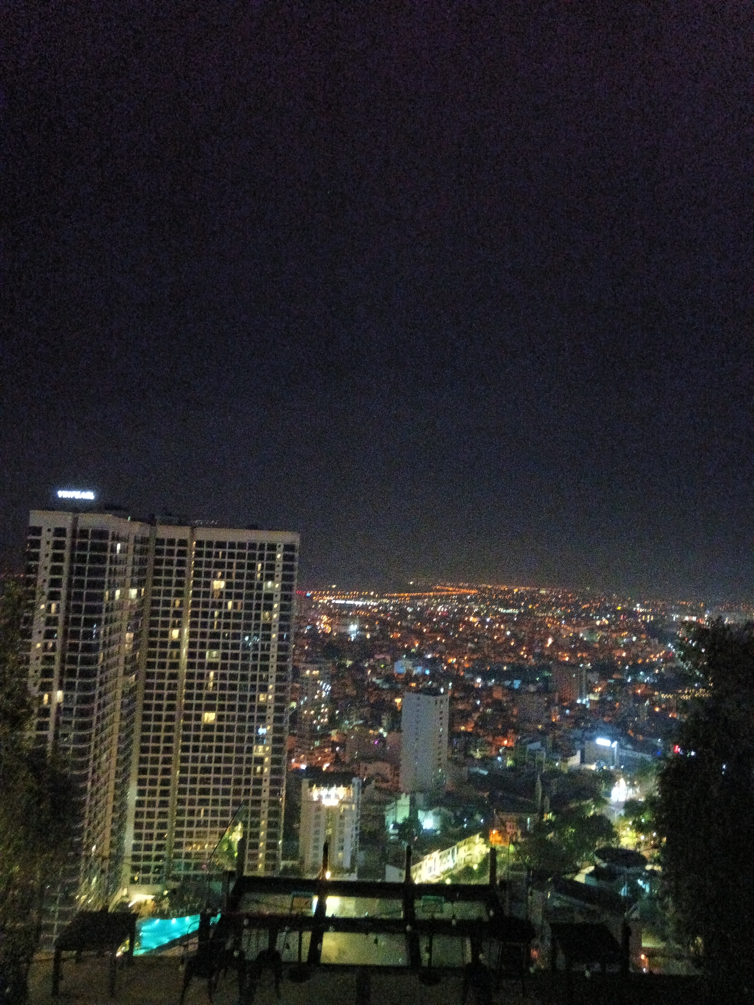 The image shows a nighttime cityscape of Nha Trang Ward, Vietnam. A tall, modern high-rise building with illuminated windows dominates the left side of the frame. A swimming pool is visible at the base of the building. In the background, a dense urban area extends towards the horizon, punctuated by numerous lights. The dark sky occupies the upper portion of the image. A partial fence or structure appears in the foreground, with some greenery partially visible on the sides of the scene.