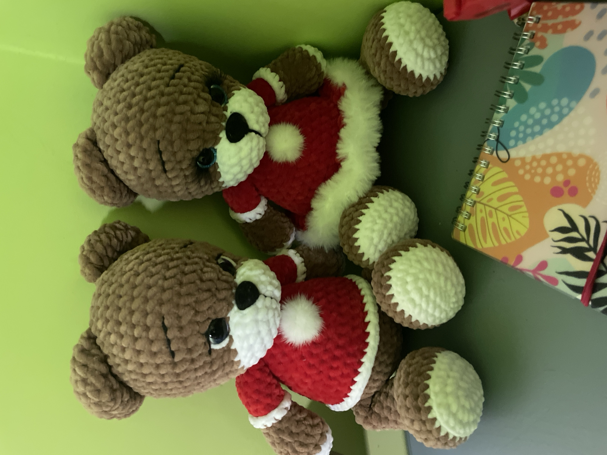 Two plush teddy bears wearing red Christmas outfits with white fur trim are lying on a green surface. The bears are made of brown yarn and have black button eyes and noses. They are positioned next to a spiral-bound notebook with a colorful, floral-like design. The location is unknown.