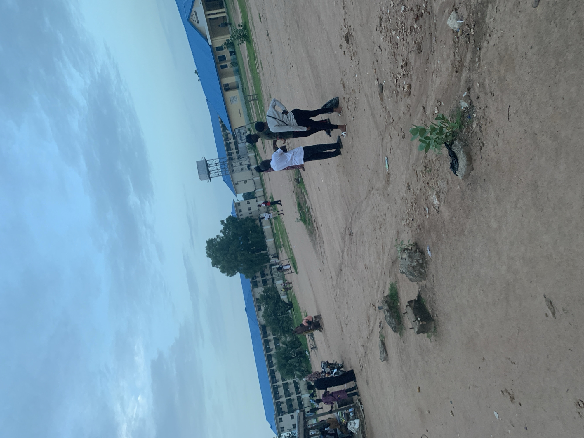 The image depicts an outdoor scene in Jalingo, Nigeria, likely a school or institutional campus based on the architecture of the buildings. In the foreground, a dirt ground area is visible with scattered rocks and vegetation. Several people are present in the scene. On the left side, a group of individuals are gathered near what appears to be a market stall with some produce displayed. In the mid-ground, three young people, two males and one female, are standing and conversing. Further in the background, more people are seen walking and standing around the buildings. The architecture features long, multi-story buildings with bright blue roofs, characteristic of public or educational institutions. A water tower is also visible behind one of the buildings. The sky is overcast with a light blue hue, suggesting it is daytime, possibly late afternoon or early morning. The overall environment appears to be a somewhat unpaved and arid area adjacent to modern structures.