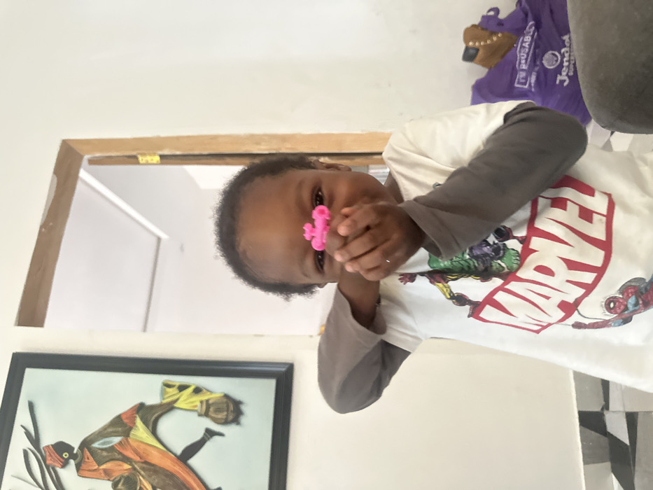 The image displays an indoor scene, consistent with a residential interior. A young child, appearing to be of African descent with dark skin and short dark hair, is positioned centrally in the foreground, looking directly at the viewer with a slight smile. The child wears a white short-sleeved t-shirt with a "MARVEL" logo and characters (Hulk, Spider-Man) printed on it, layered over a grey long-sleeved shirt. Both hands are raised to face level, holding a small, irregular pink toy object near the child's nose.
The background features white walls. To the left, a black-framed piece of wall art hangs, depicting a stylized figure in traditional attire carrying a pot, rendered in orange, brown, and yellow tones. To the right, an open wooden door frame with a visible gold-colored hinge leads into another brightly lit room with white walls and a light-colored ceiling featuring a subtle molding detail. In the lower right background, a purple bag with partial text "Jendol SUPERM..." and "REUSABLE" is visible, along with a partial brown boot. The floor in the lower left foreground shows a pattern of light and dark tiles. The location is an indoor space, and while context suggests a residence in Ilishan, Nigeria, no specific visual markers within the image confirm this city.