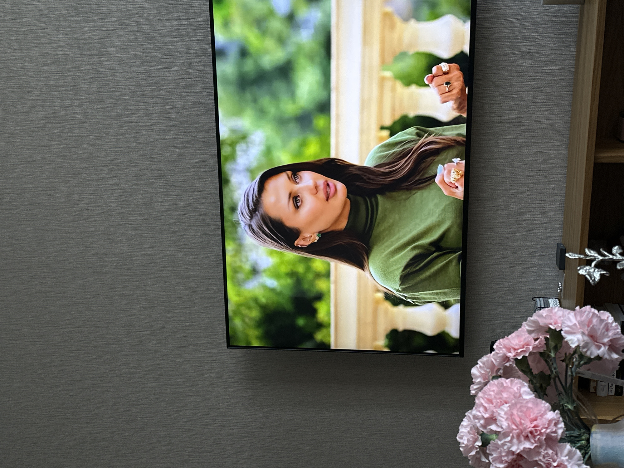 The image shows a television screen displaying a woman with long brown hair wearing a green turtleneck. She is outdoors in a bright, possibly garden setting. The television is mounted on a gray wall, with a wooden shelf unit to the right. A bouquet of pink carnations is partially obscuring the lower right corner of the screen and part of the shelf. The scene appears to be indoors, possibly in a living room. There is no discernible information to determine the location in Yerevan, Armenia.